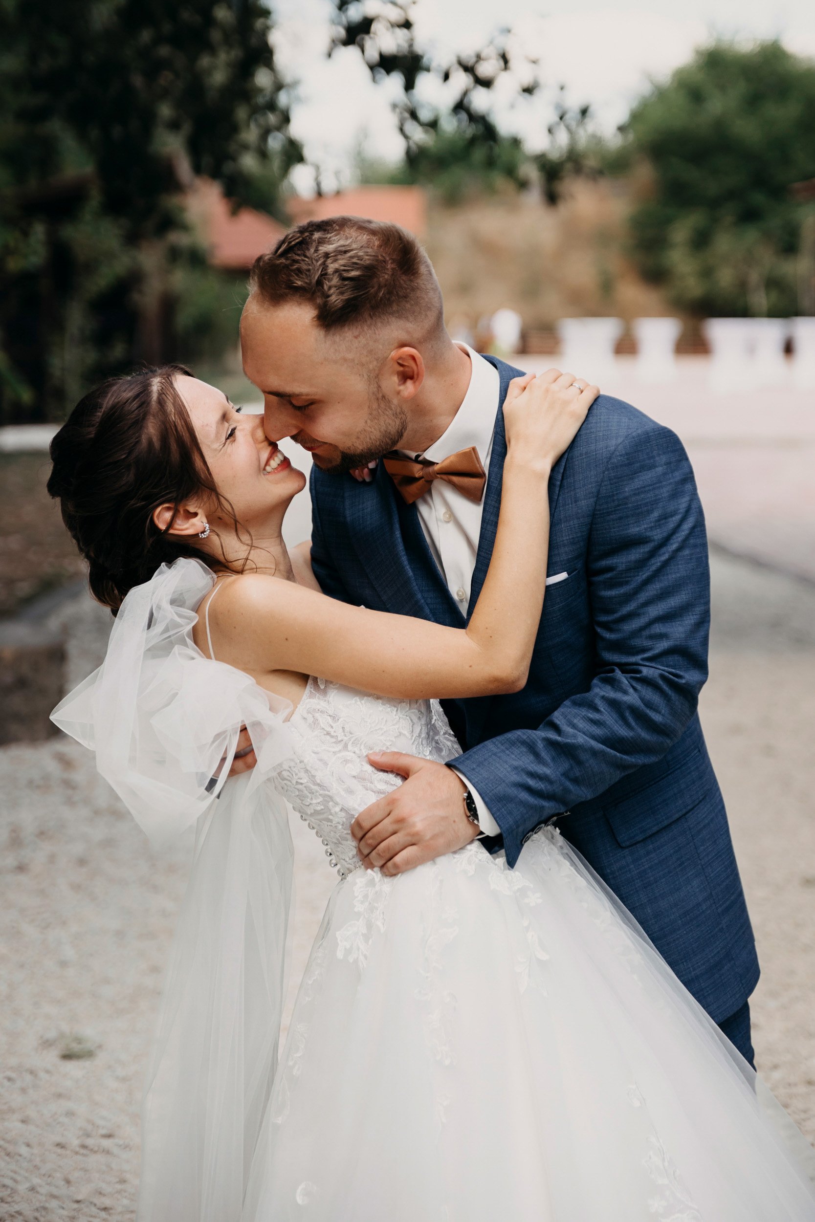 A bride and groom dancing outdoors, gazing into each other's eyes with smiles, surrounded by trees and a stone walkway.