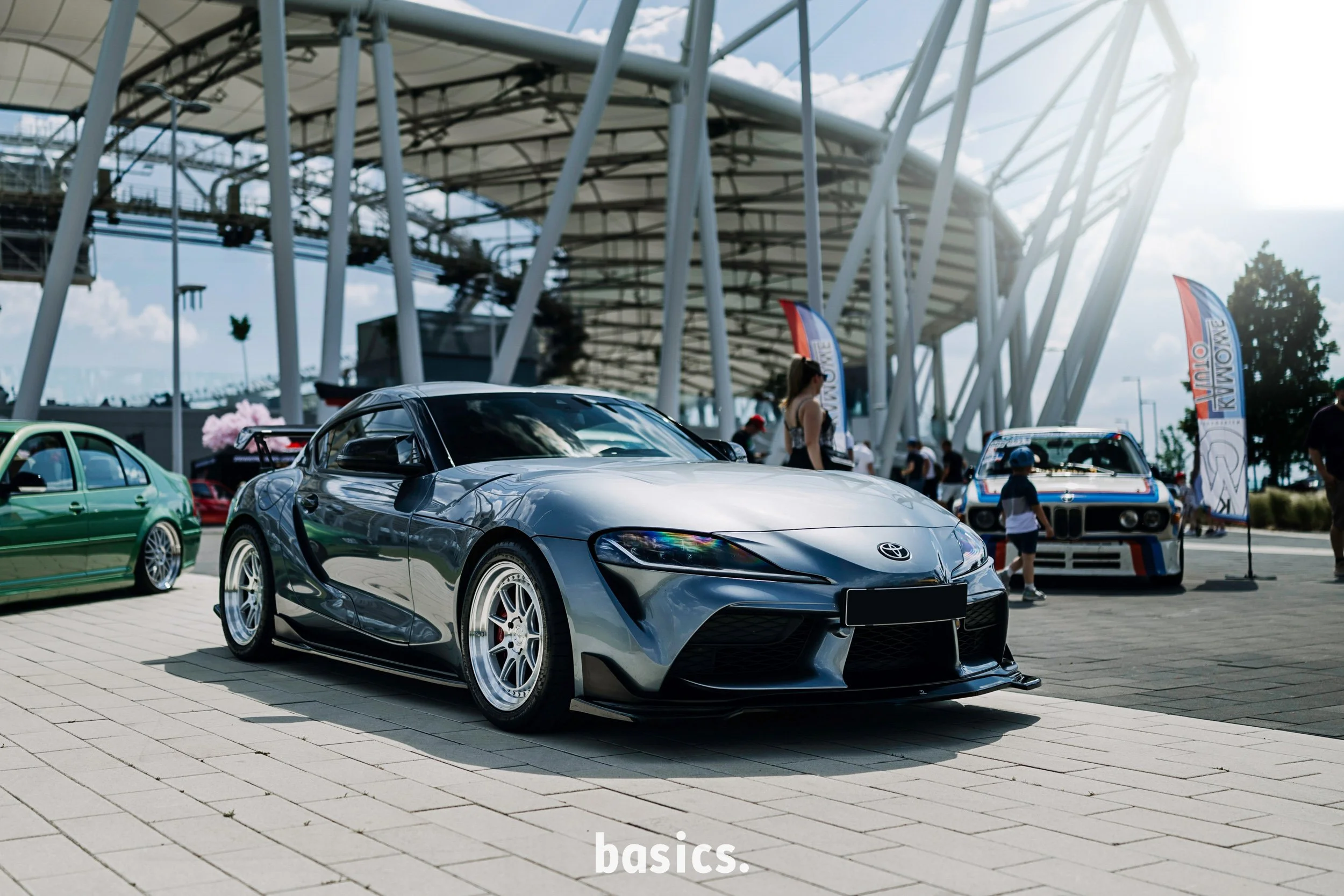 A silver sports car is parked outside a modern building with a glass and steel roof structure. There are other colorful cars and people in the background at what appears to be a car show.