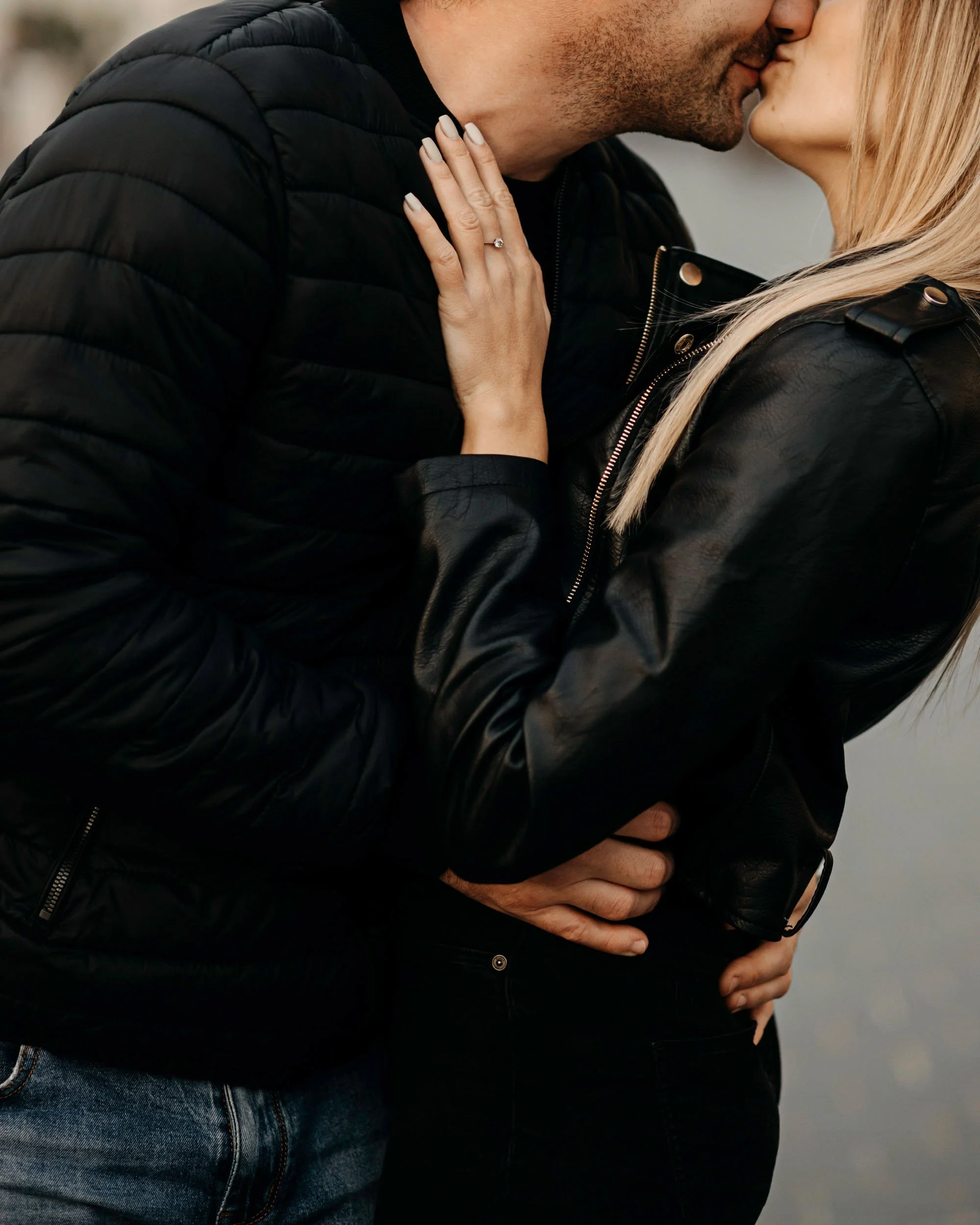 A close-up of a couple kissing, with the woman resting her hand on the man's shoulder, displaying an engagement ring, both wearing black jackets.