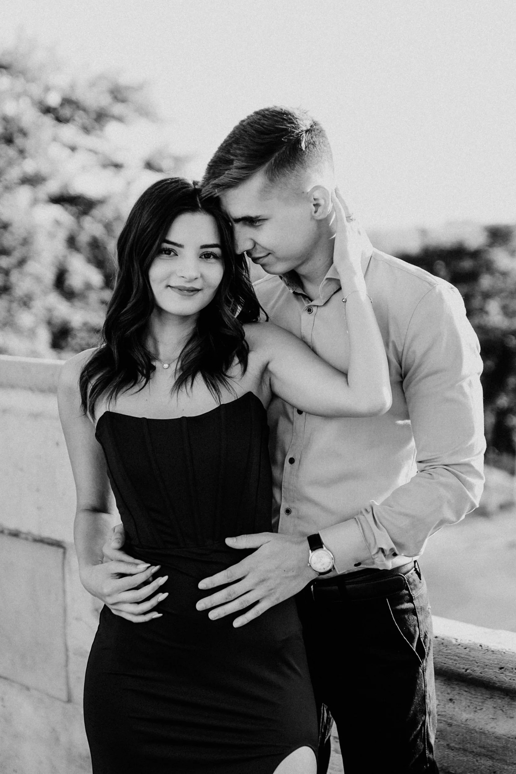 A black and white photograph of a young couple embracing outdoors. The woman is smiling at the camera, while the man gently rests his face against her forehead. The woman's hand is behind her head, and the man's hand is on her waist. blurred trees ar