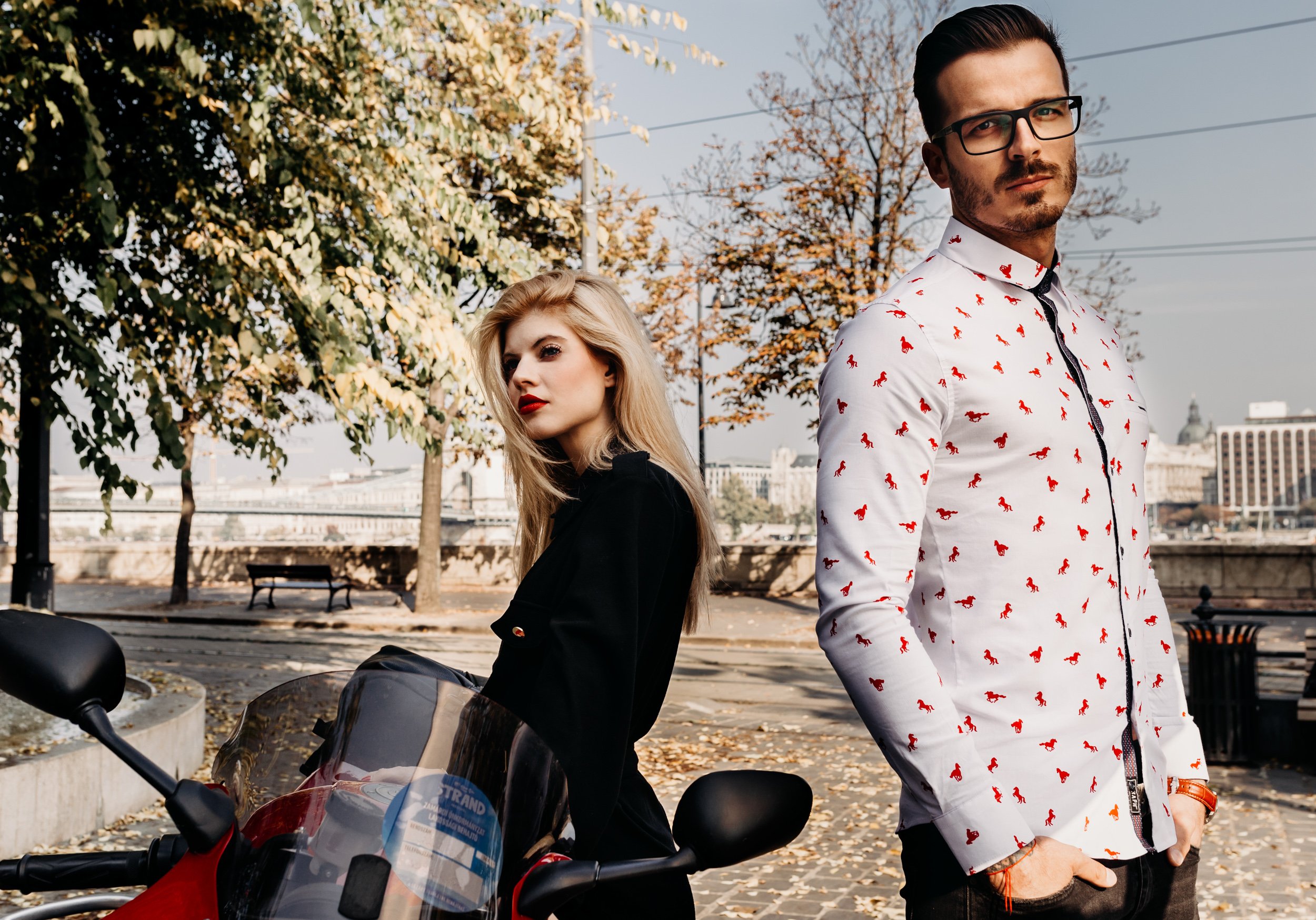 A man wearing glasses and a white shirt with red horse patterns is standing outdoors during autumn. A woman with long blonde hair and red lipstick is sitting on a motorcycle next to him, with trees and city buildings in the background.
