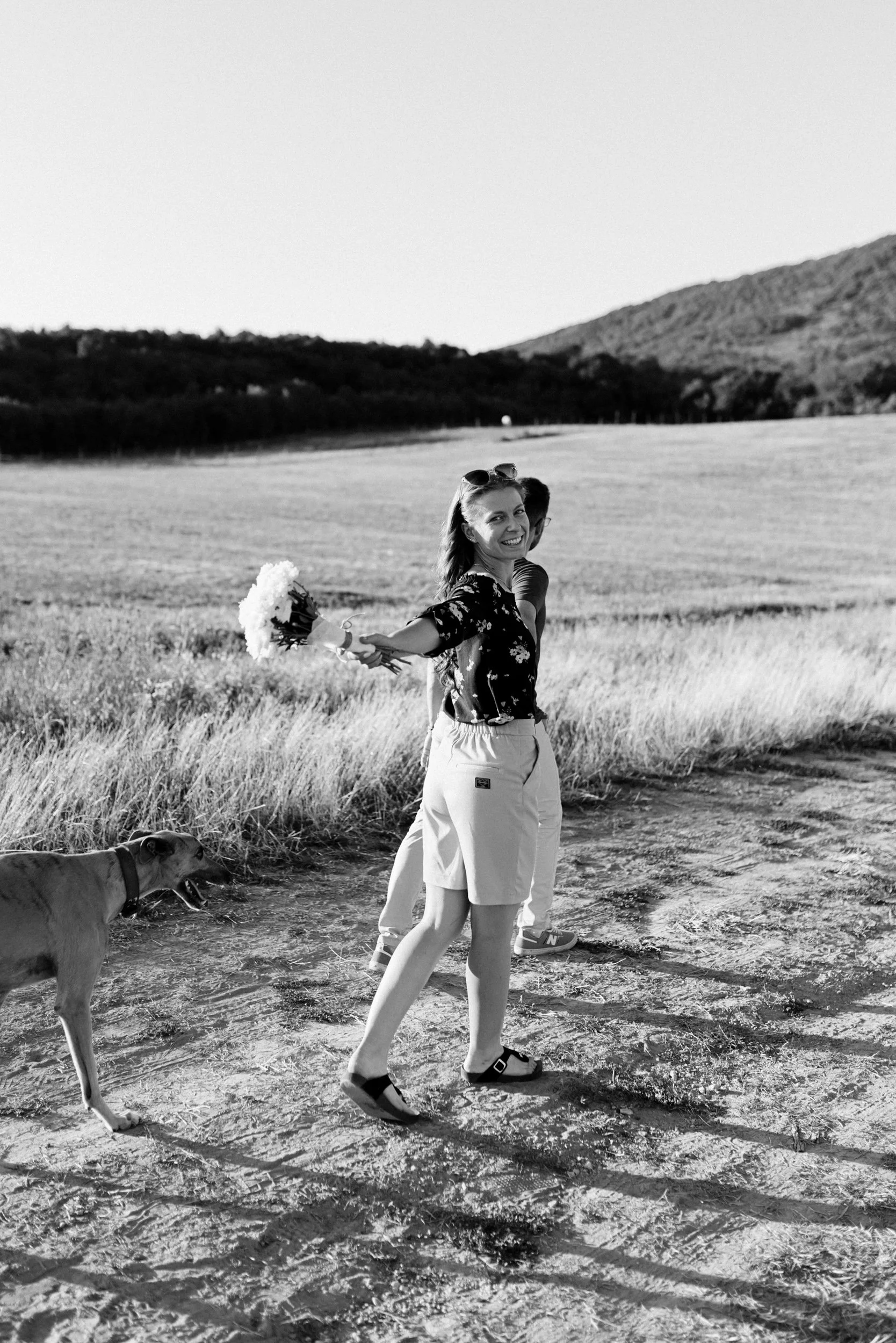 A woman smiling in a black floral shirt and light-colored shorts walks on a dirt path in an open field with a dog on a leash. She has sunglasses on her head and is holding a bouquet of flowers. In the background, another person and a scenic landscape