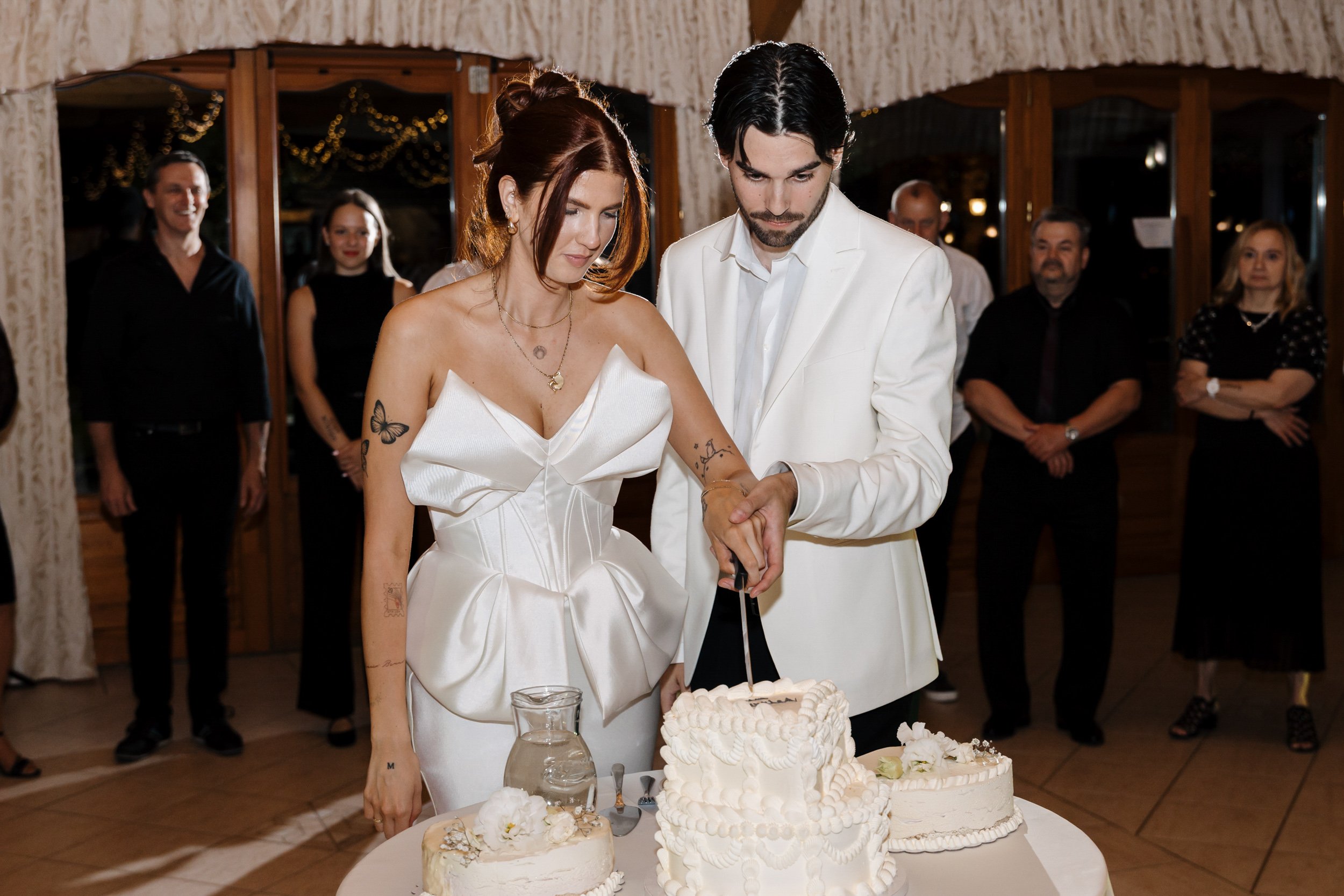 A couple dressed in wedding attire cutting a wedding cake at their reception, with guests watching in the background.