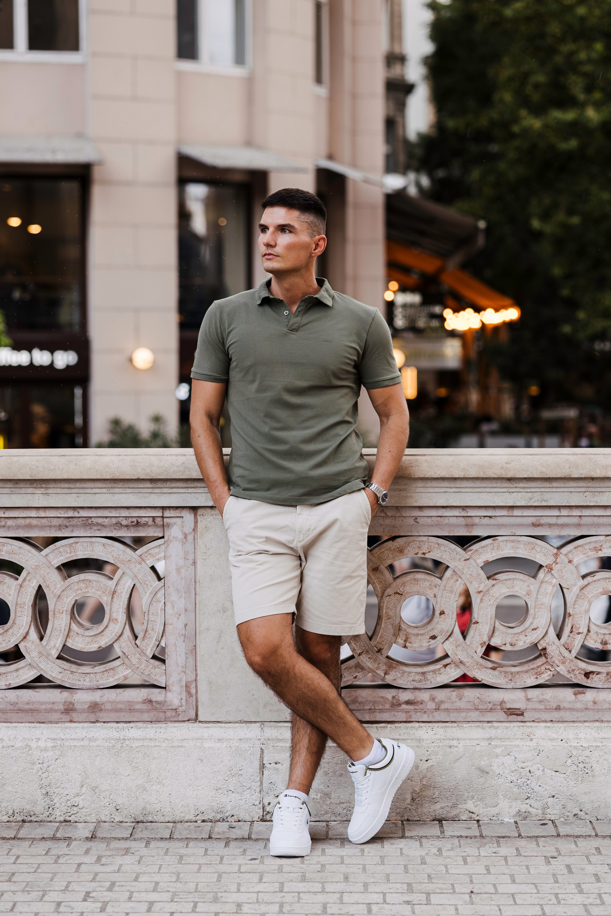 A man wearing a green polo shirt, beige shorts, white sneakers, and a watch leans against a decorative stone railing in front of a city street with buildings and trees during the daytime.