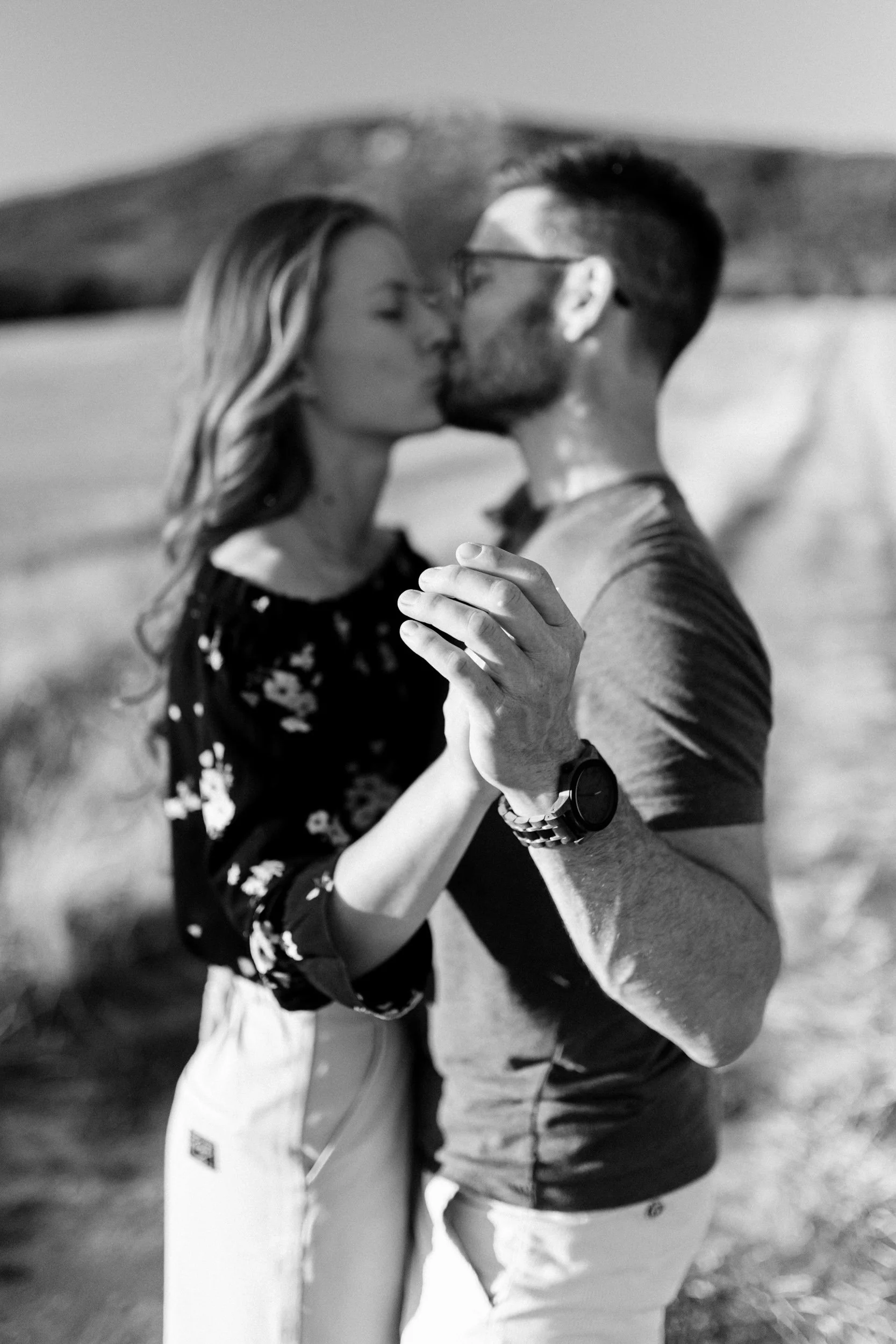 A man and woman share a kiss outdoors, with the woman gently holding the man's arm. The image is in black and white, and they are dressed casually.