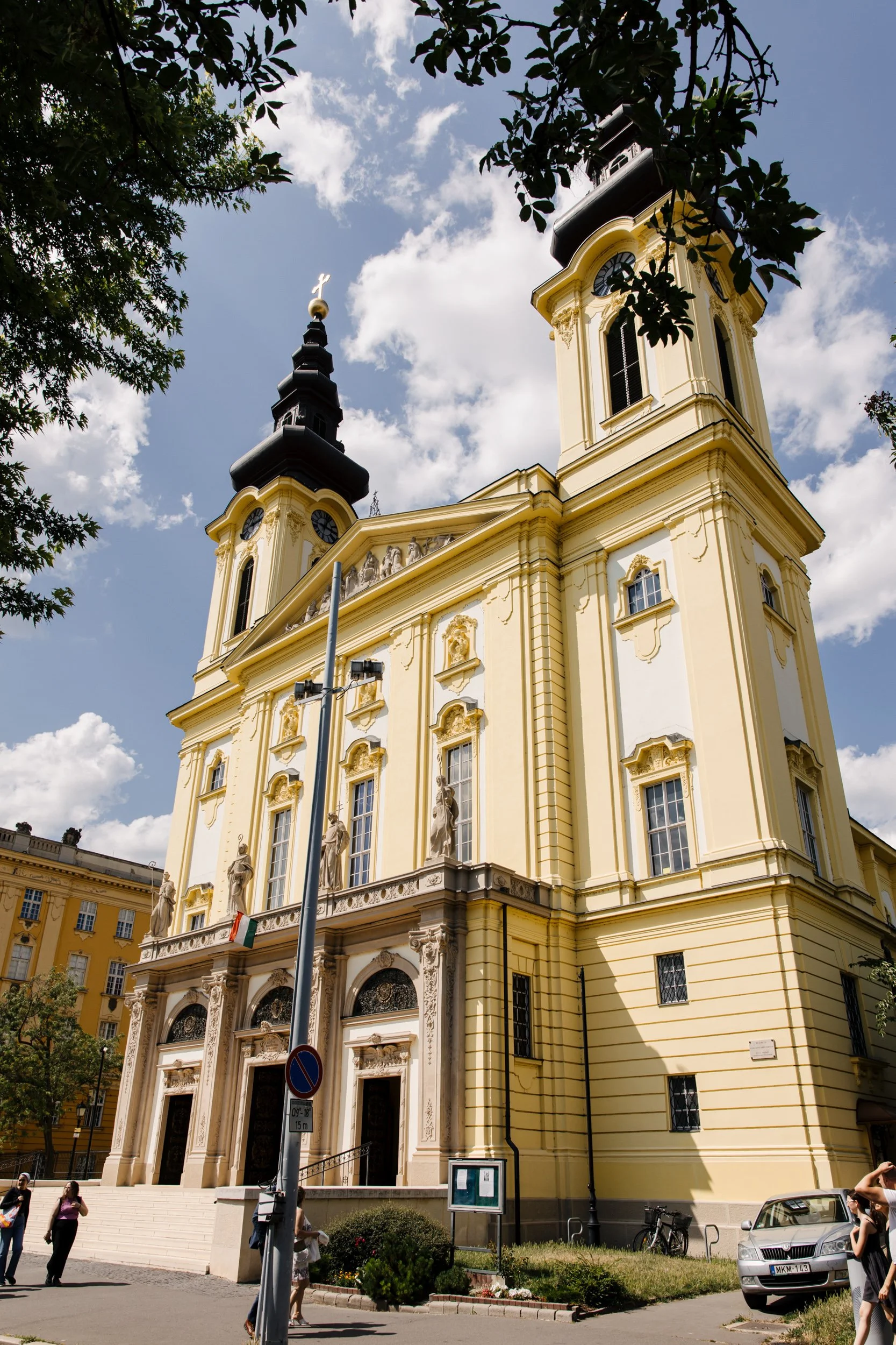 A yellow baroque-style church with twin clock towers topped with black onion domes and crosses, surrounded by trees. Several people are walking in front, and parked cars can be seen along the street.