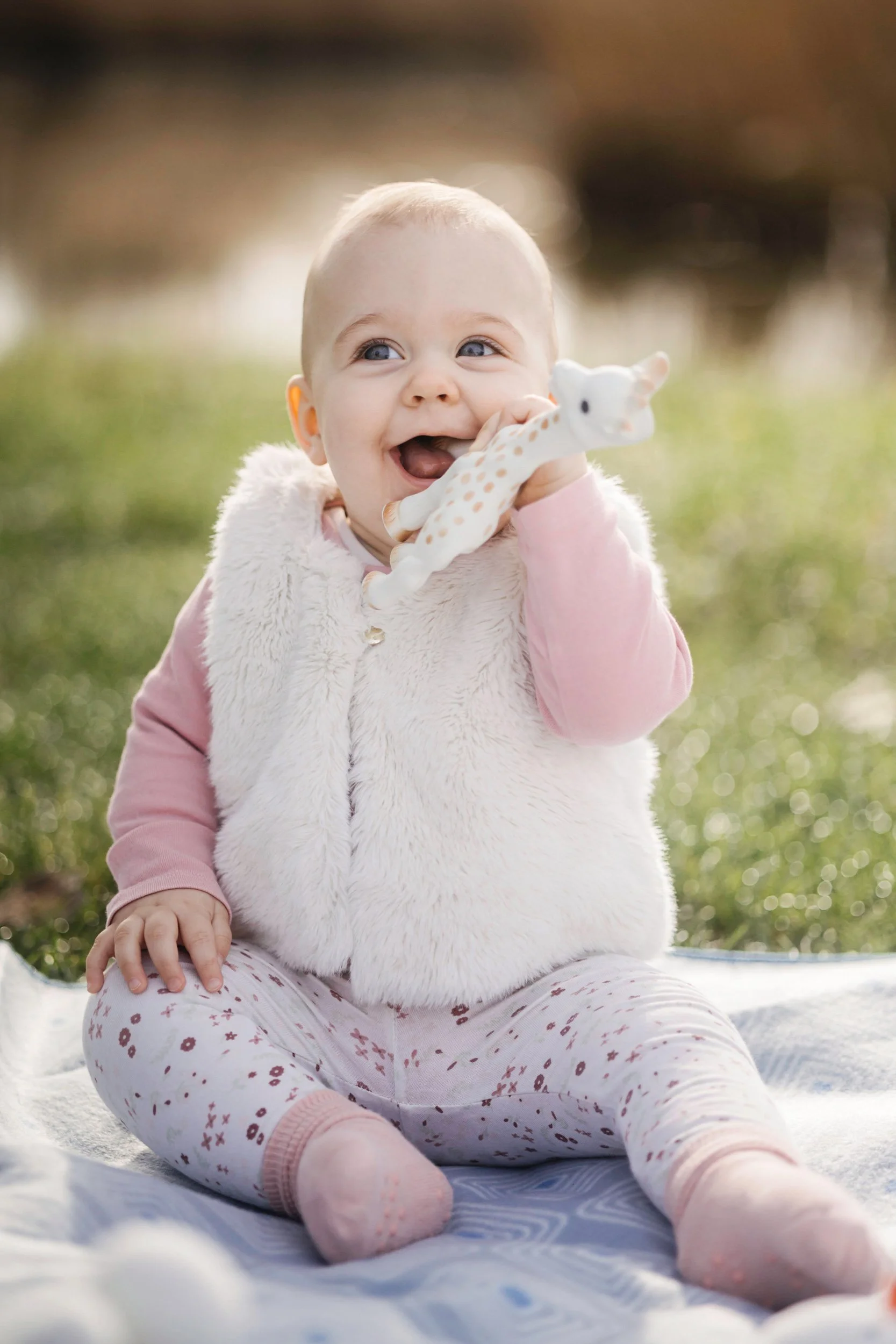 A smiling baby girl with blue eyes, wearing a pink long-sleeve shirt, a white fuzzy vest, and pink and white patterned leggings, sitting outdoors on a blanket in a grassy area, holding a soft giraffe toy.