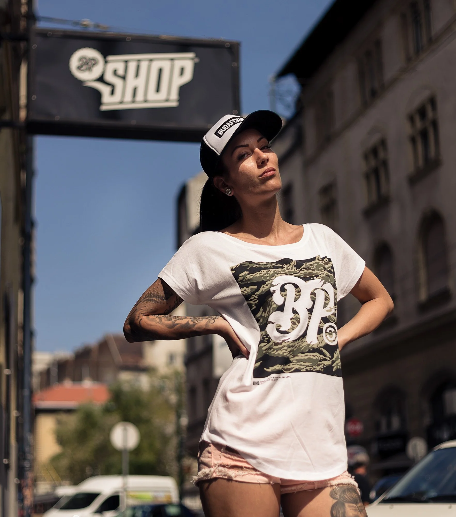 A woman with dark hair and tattoos on her left arm and right thigh standing outdoors in front of buildings and cars, wearing a white T-shirt with a graphic and a black and white cap, looking slightly to the side.