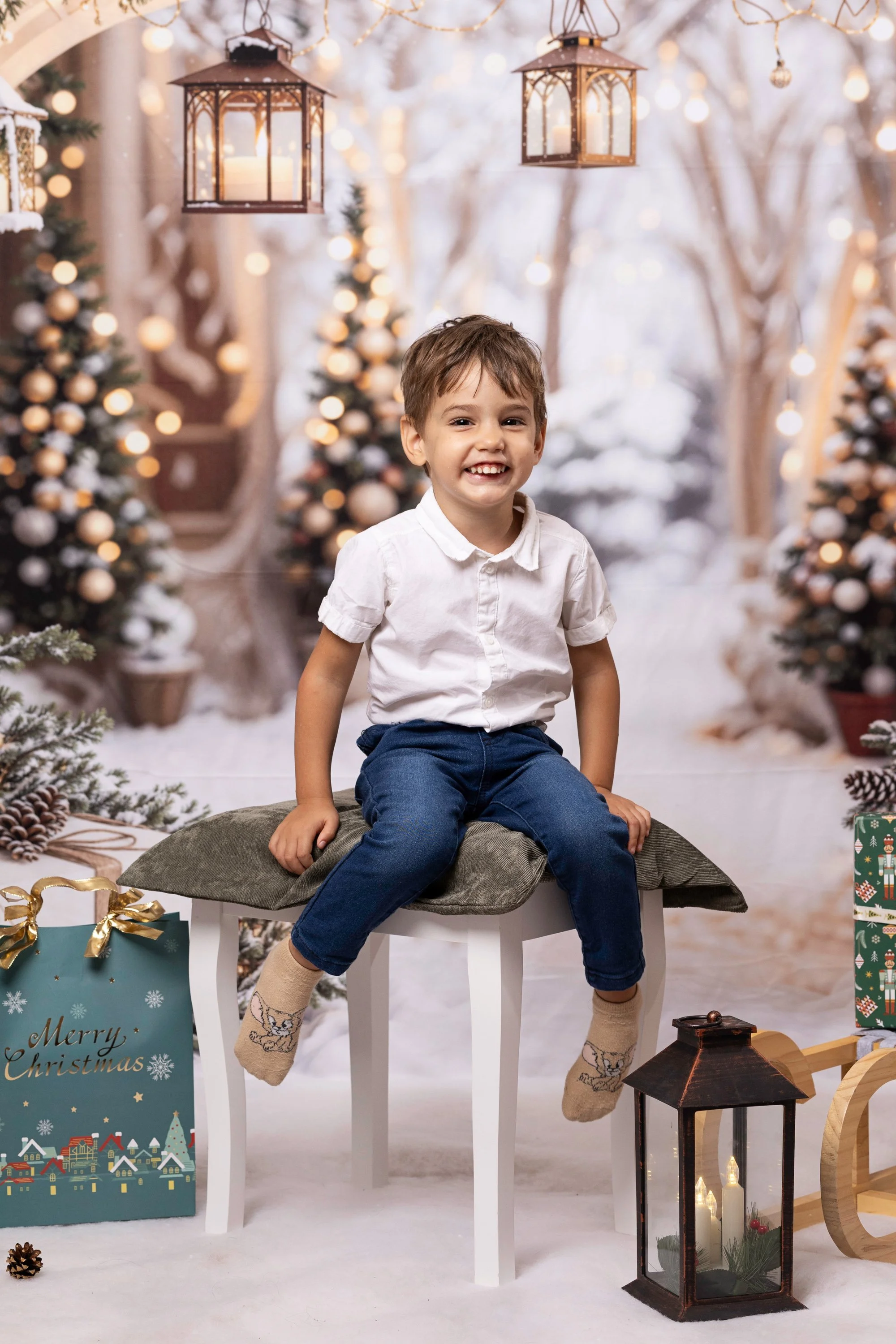 A young boy sitting on a cushioned stool in festive Christmas decor, surrounded by wrapped gifts, holiday decorations, and Christmas trees with ornaments, in a winter-themed setting with hanging lanterns and warm lighting.