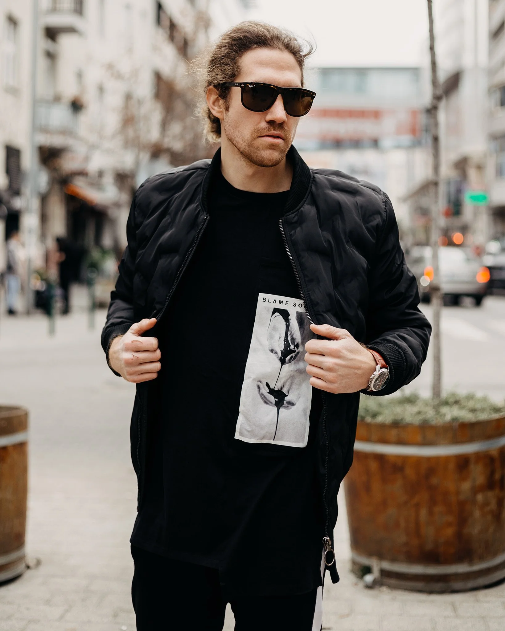 A man with long hair, wearing sunglasses, a black jacket, and a black t-shirt with a graphic, standing outdoors in what appears to be an urban setting with buildings and trees in the background.