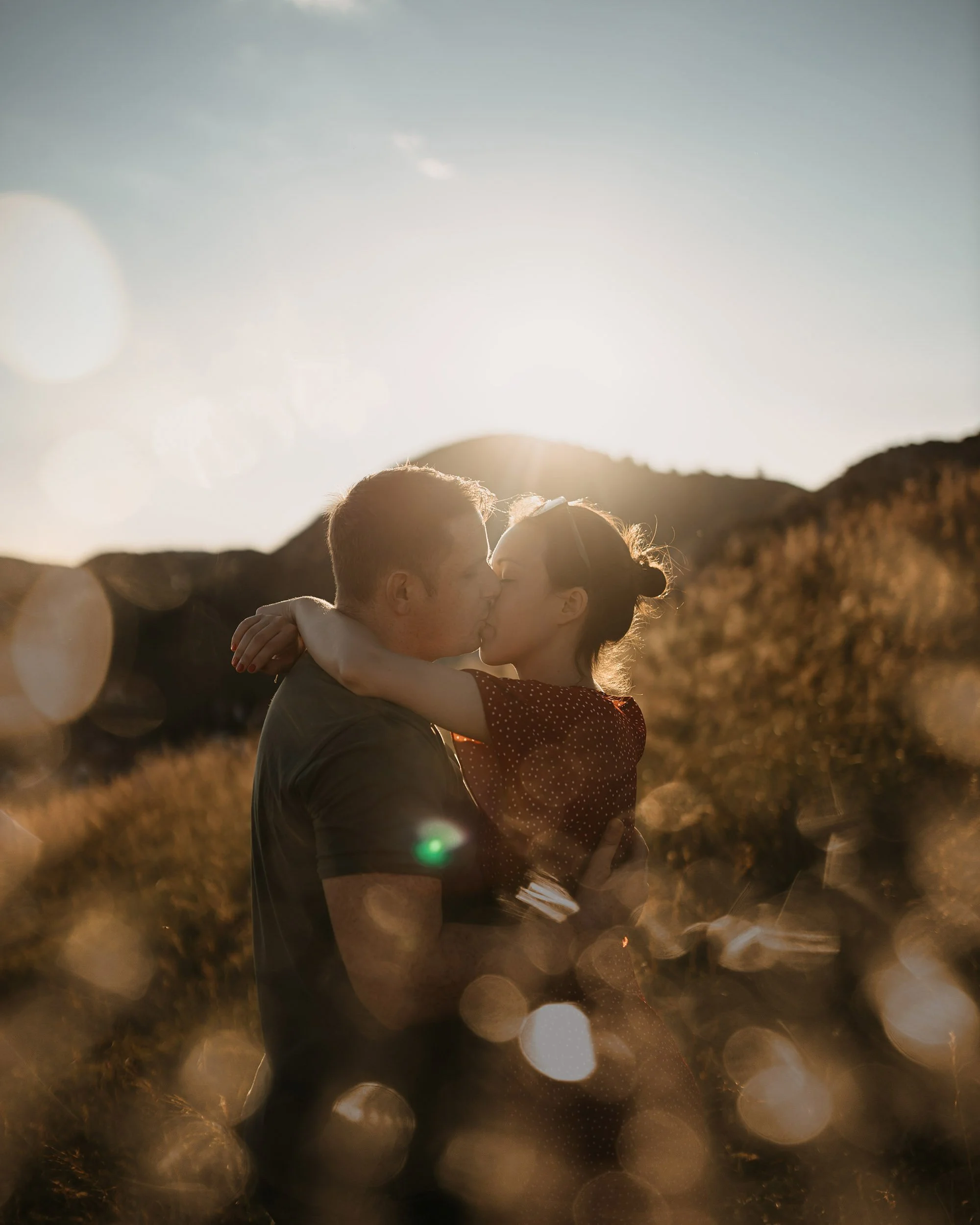 A couple sharing a kiss in a scenic outdoor setting during sunset with bokeh lights and mountainous background.