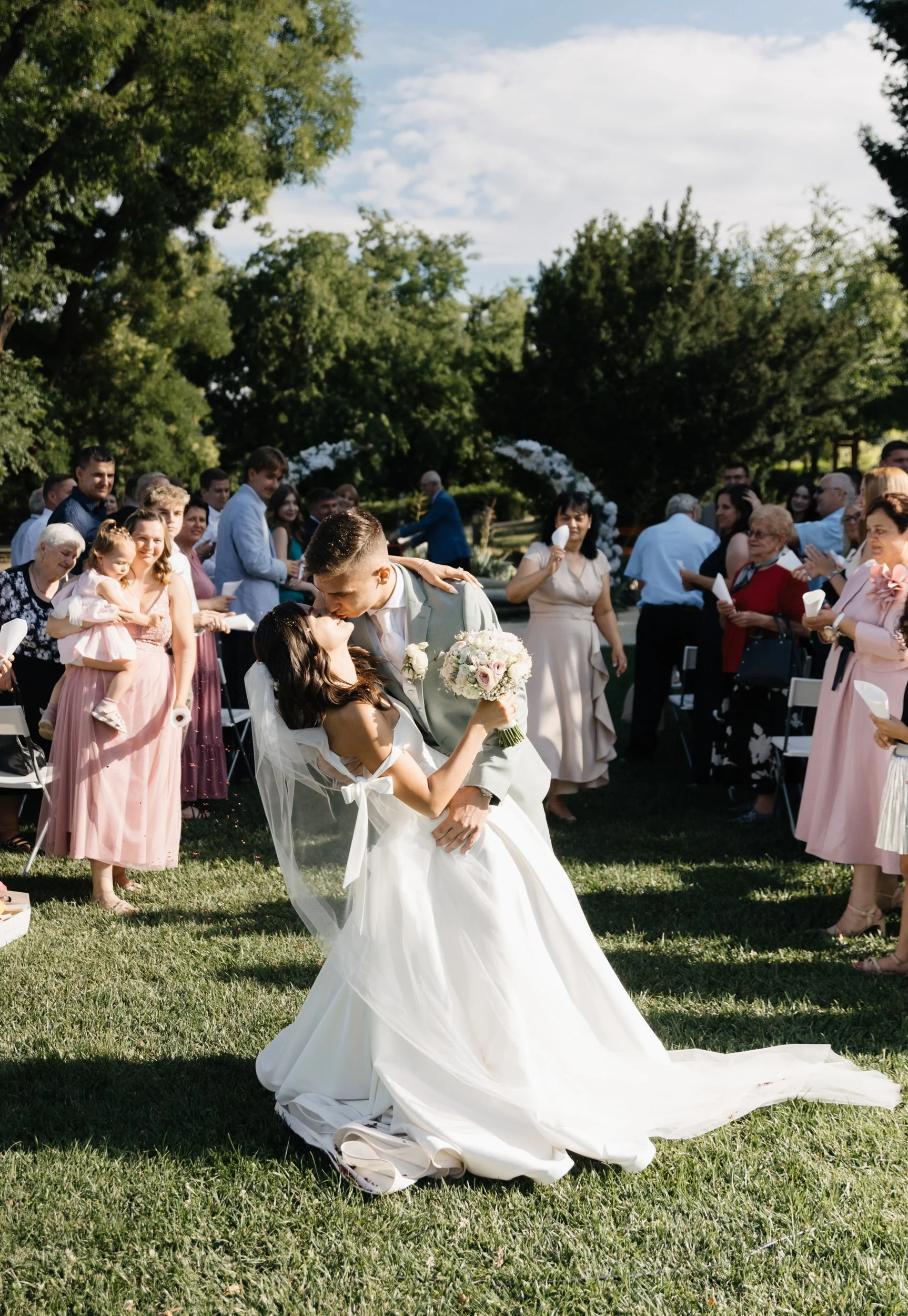 A wedding scene outdoors with a bride and groom kissing, surrounded by guests. The bride, wearing a white wedding dress, is holding a bouquet of flowers. The groom is dressed in a light gray suit. The guests observe, some smiling and holding programs