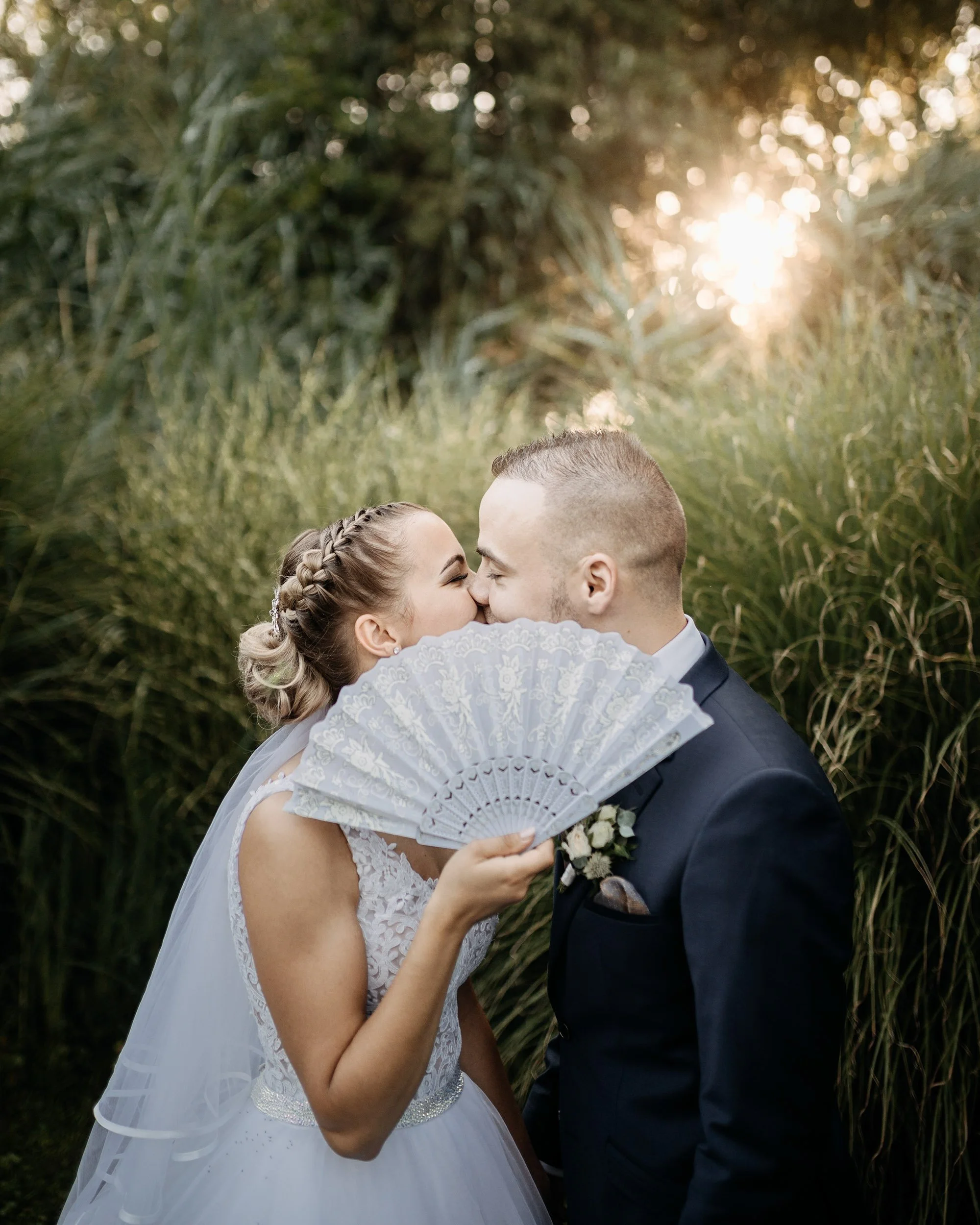 A bride and groom kissing outdoors during their wedding, with a grassy background and sunlight filtering through trees.