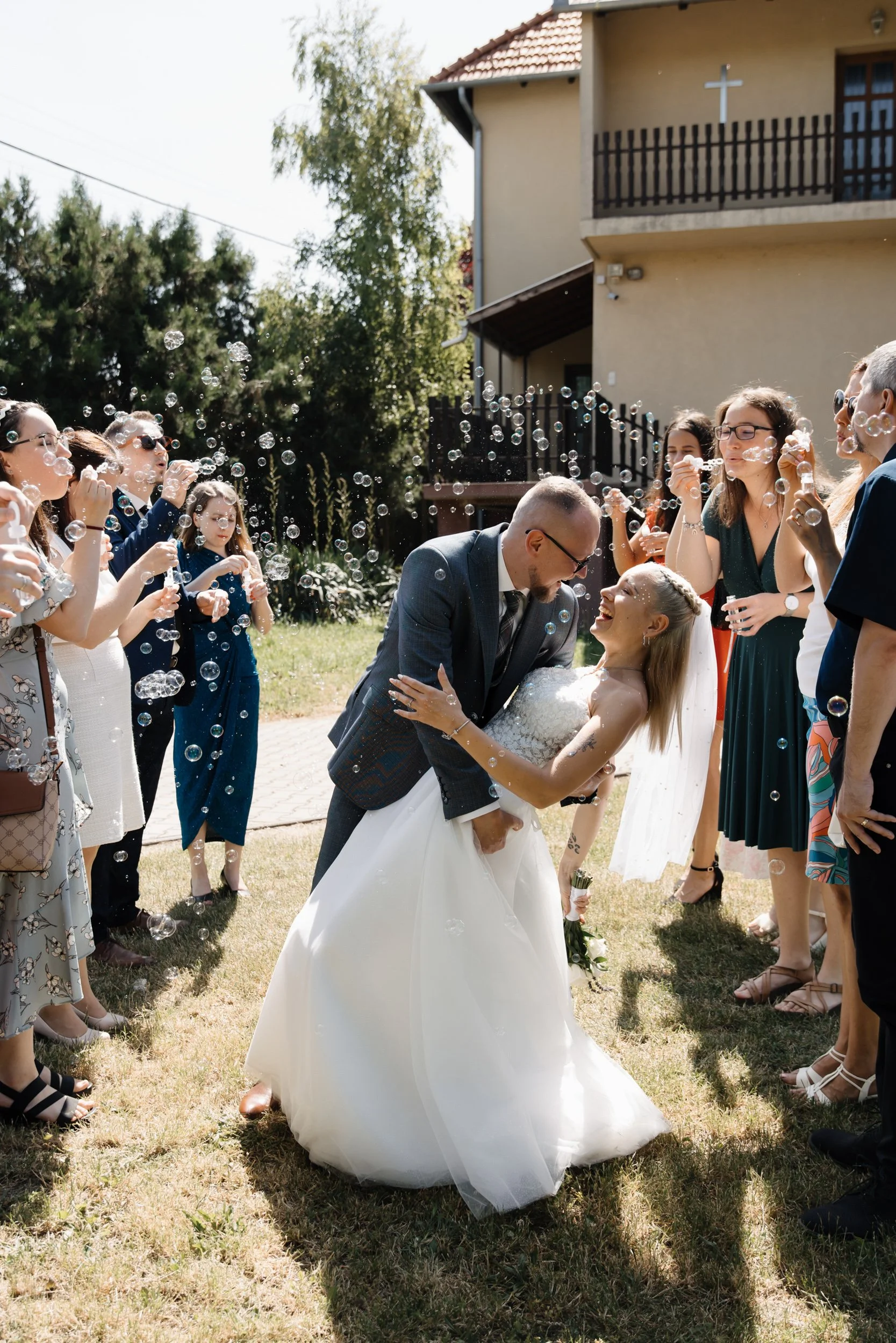 A newlywed couple is surrounded by friends and family blowing bubbles during an outdoor wedding celebration.