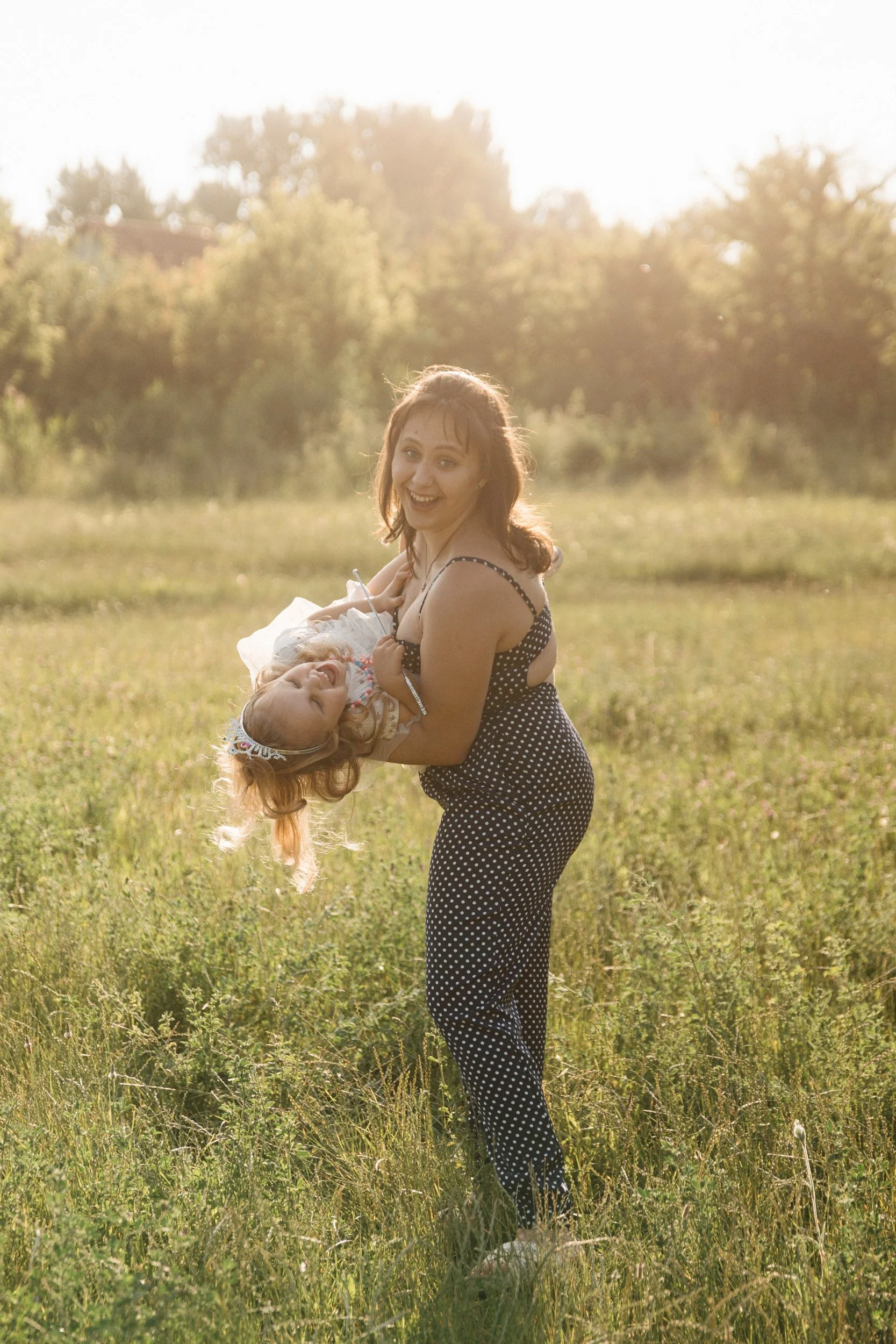 A woman holding a joyous young girl in a grassy field during sunset, both smiling and enjoying each other's company.