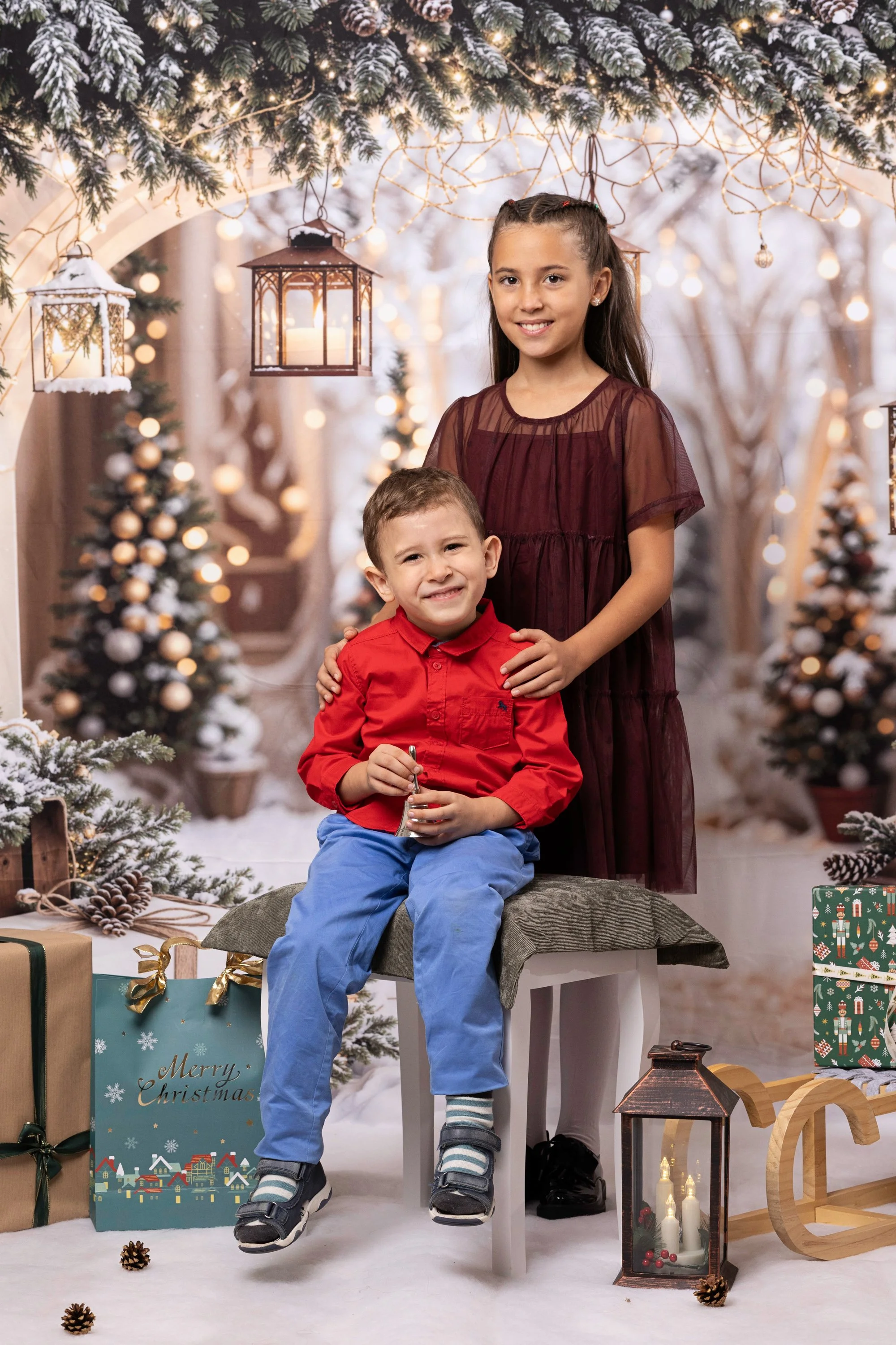 A girl and a boy posing in a Christmas-themed setting with decorated trees, wrapped presents, and festive lights.