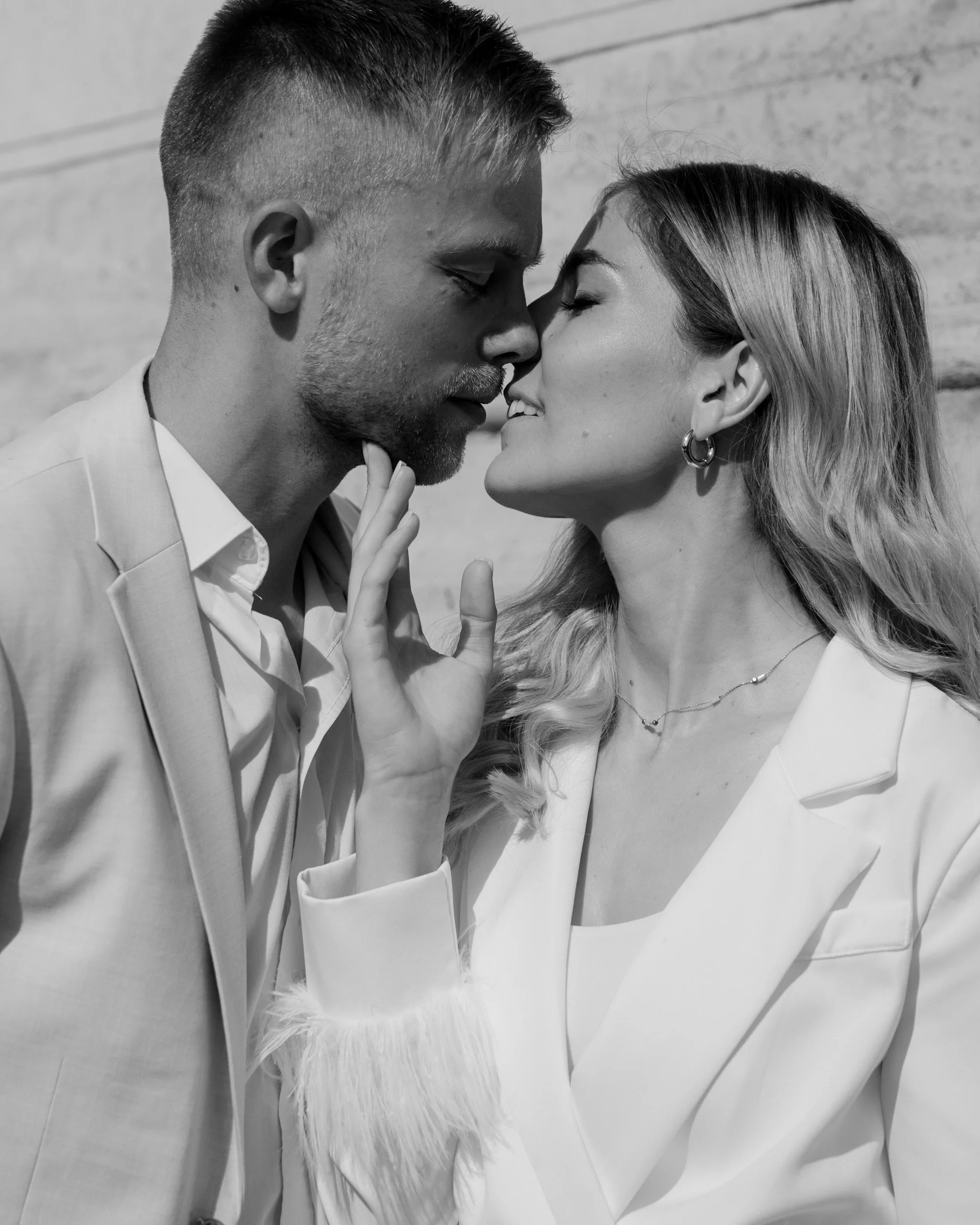 A black and white photo of a young man and woman standing very close, with their foreheads touching and eyes closed. The man has short hair and a beard, and is wearing a light-colored suit. The woman has long, wavy hair, earrings, and a necklace, and