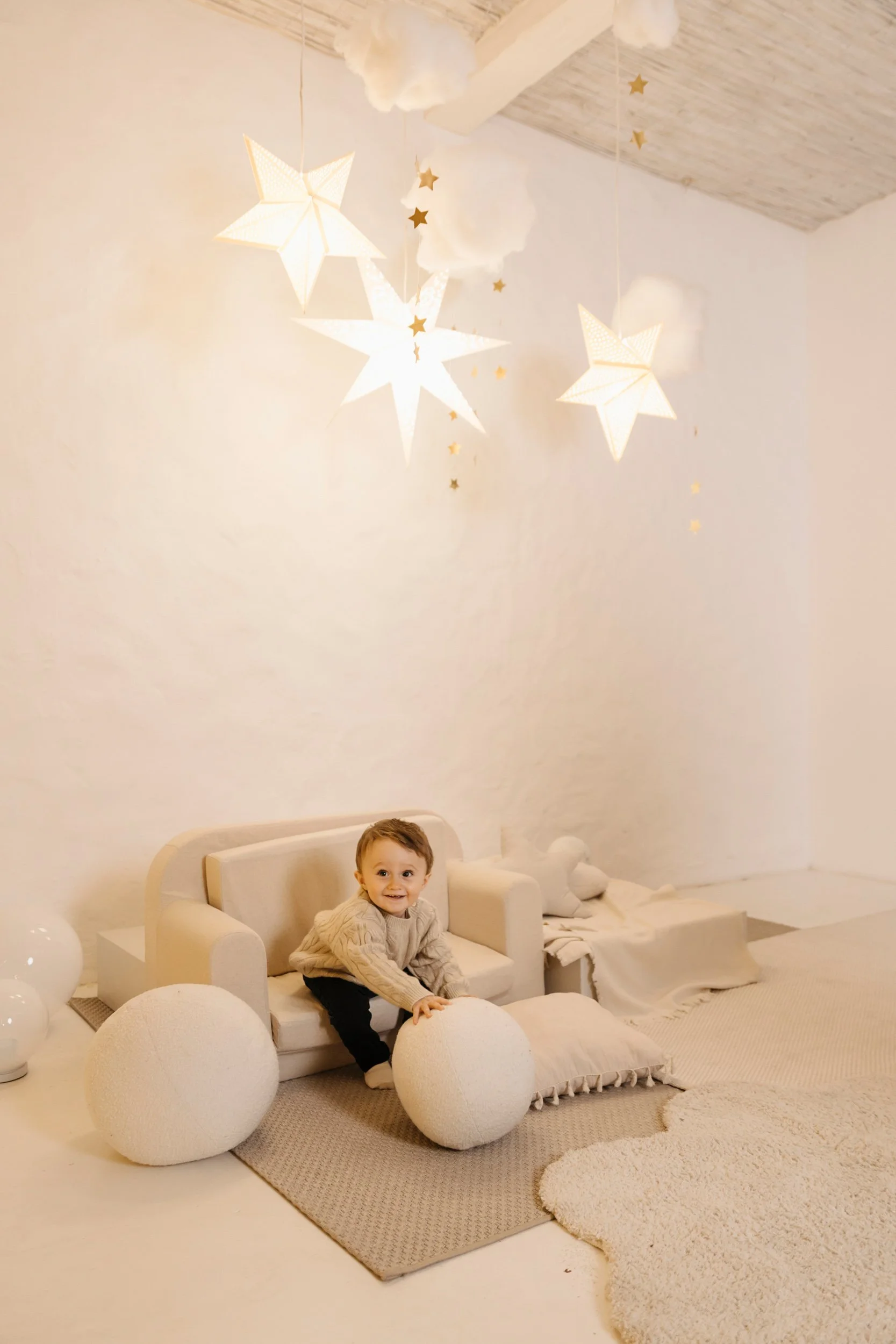 A young child sitting on a beige toy couch in a room decorated with hanging paper stars and clouds, plush clouds on the floor, soft area rugs, and star-shaped paper lanterns in the ceiling.