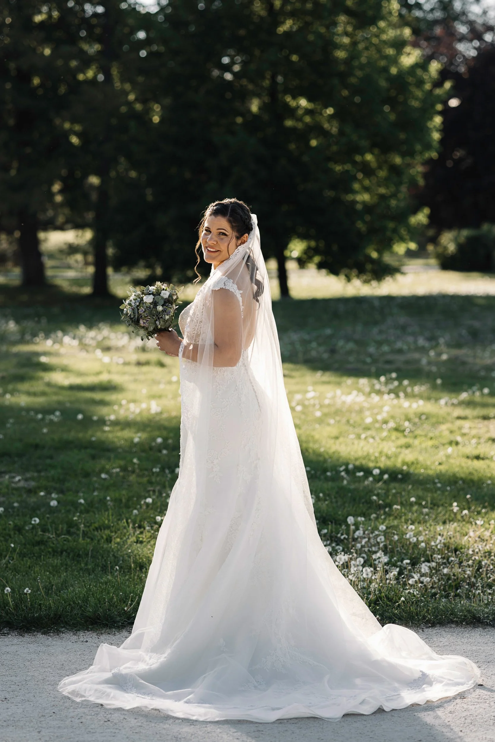 A smiling bride in a white wedding dress with lace details, holding a bouquet, standing outdoors in a park with lush green grass and trees in the background during daytime.