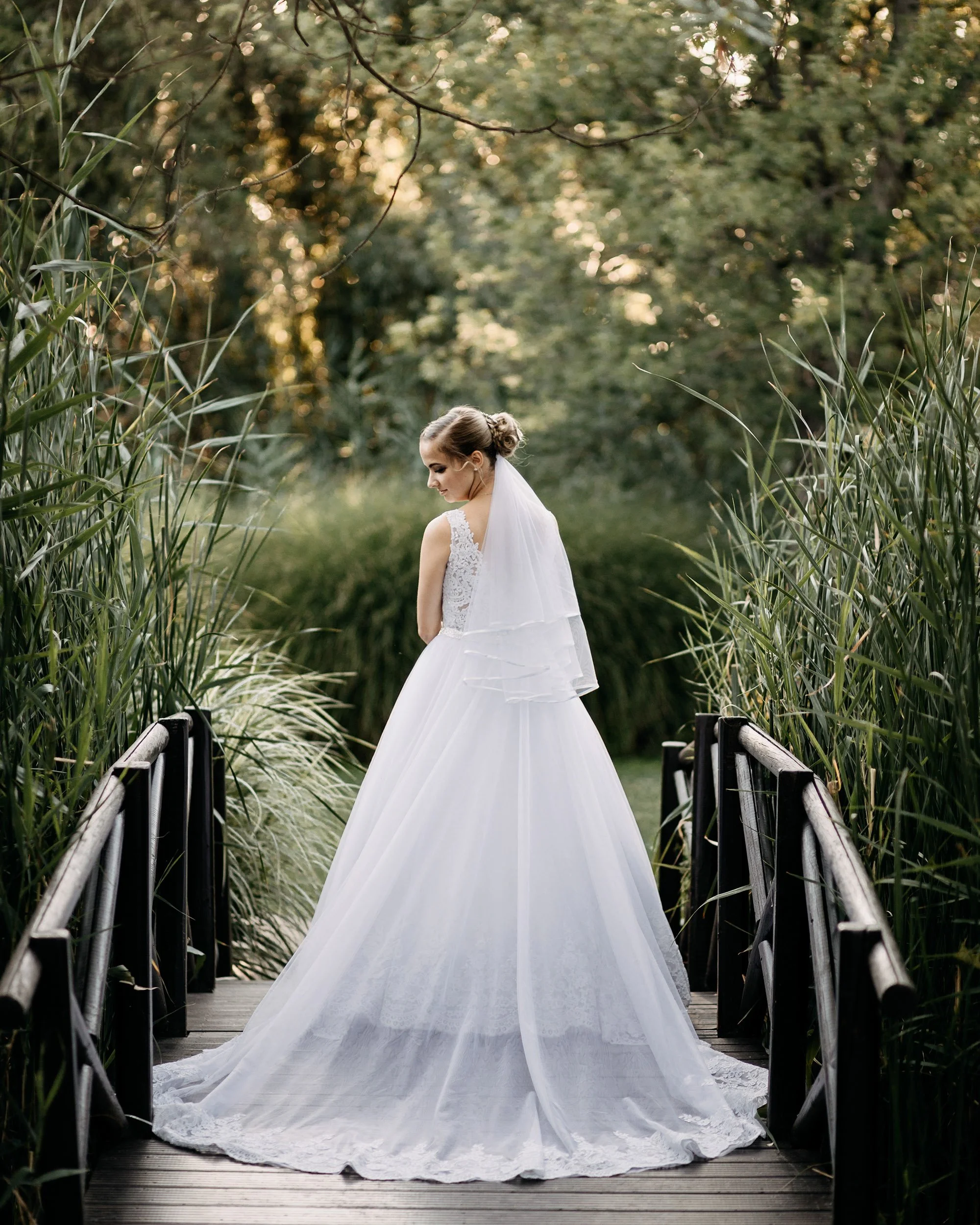 A bride in a white wedding gown with lace details and a long veil standing on a wooden bridge surrounded by tall green grass and trees, with sunlight filtering through the branches.