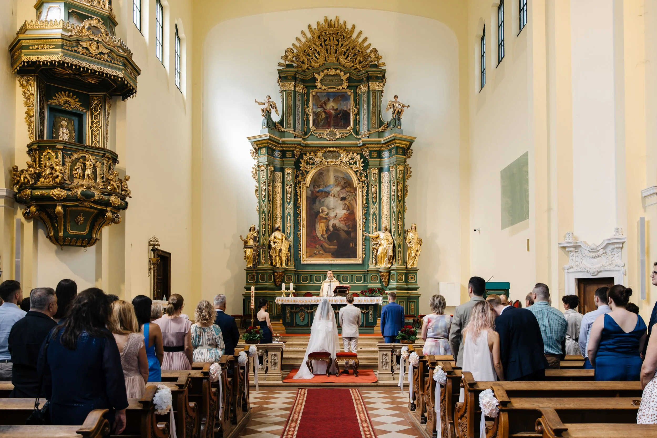 A congregation gathered in a church during a wedding ceremony with the bride walking down the aisle towards the altar, which is decorated with religious paintings, gold accents, and statues, under a large ornate altarpiece.