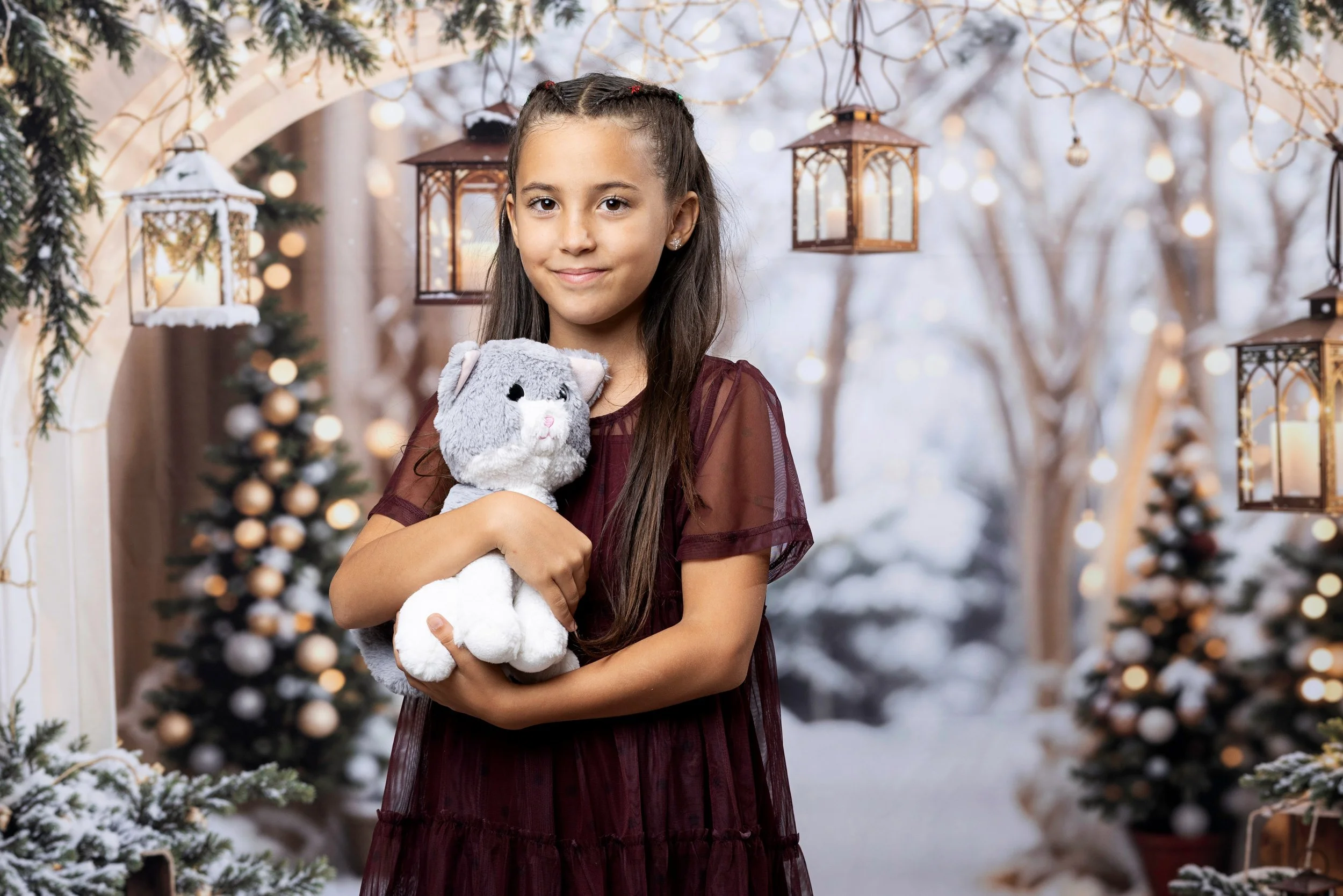A young girl with long brown hair in braids, wearing a burgundy dress, is holding a plush gray mouse toy. She is standing outdoors in a snowy scene decorated with Christmas ornaments, with lit lanterns hanging from above and Christmas trees in the ba