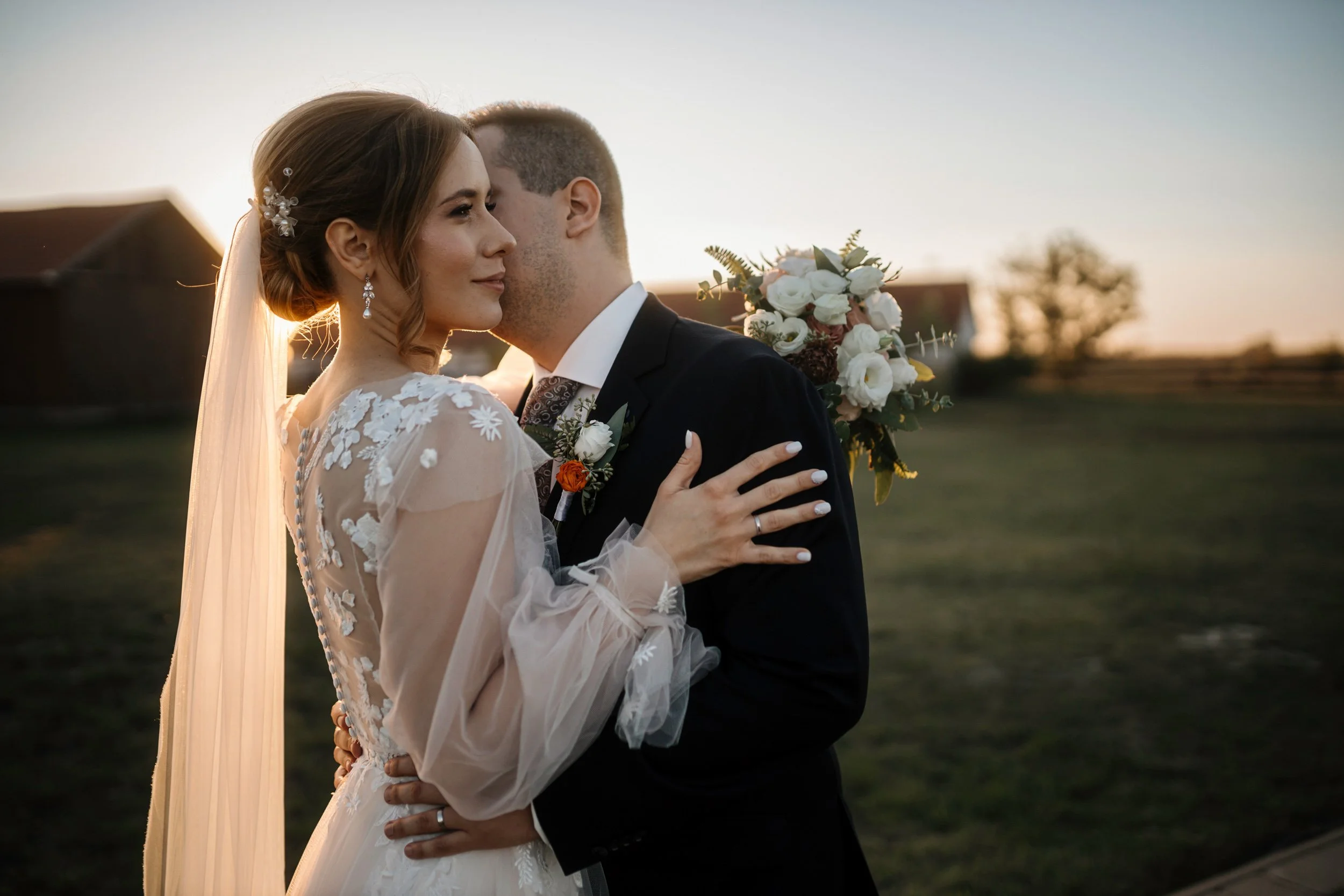 A bride and groom embrace during their outdoor wedding at sunset. The bride wears a lace wedding dress with floral embroidery and a pearl hair accessory, while the groom wears a black suit with a boutonniere. The bride's hand rests on the groom's sho