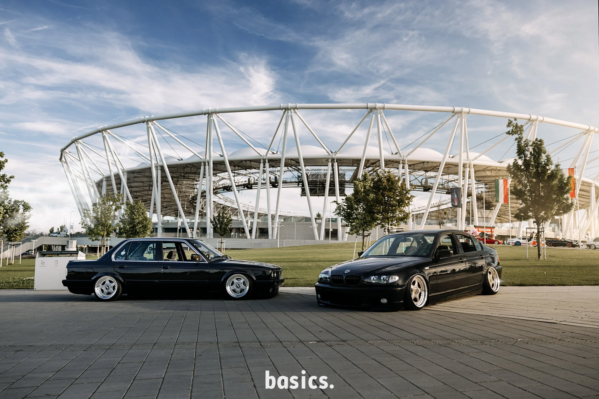 A parking lot with two black BMW cars parked on a paved surface, with trees and a large modern stadium in the background and partly cloudy sky overhead.