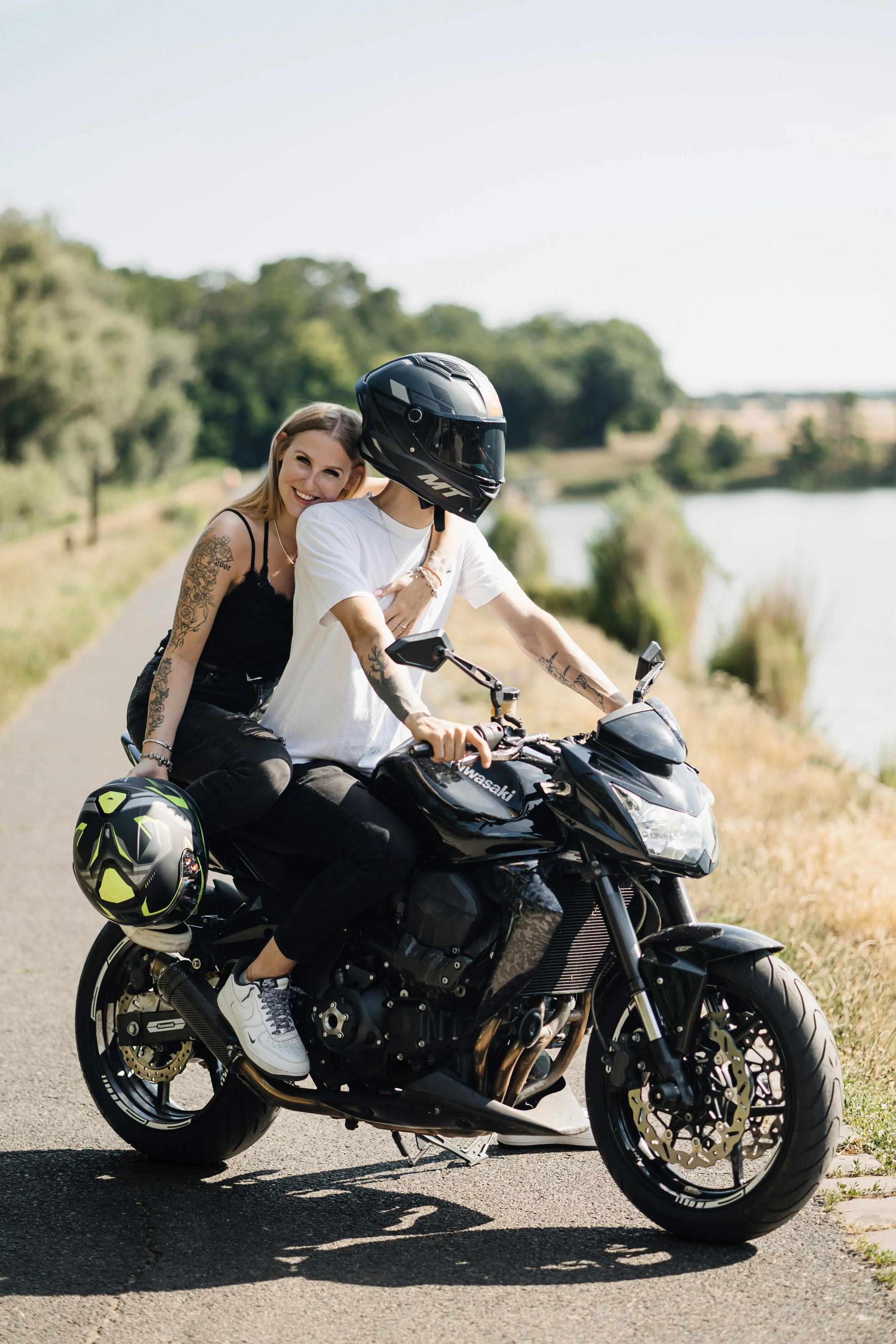 A young woman with tattoos on her arm, dressed in a black tank top and black pants, smiling and sitting behind a person wearing a motorcycle helmet and white t-shirt, riding a black Kawasaki motorcycle along a scenic road near a body of water with tr
