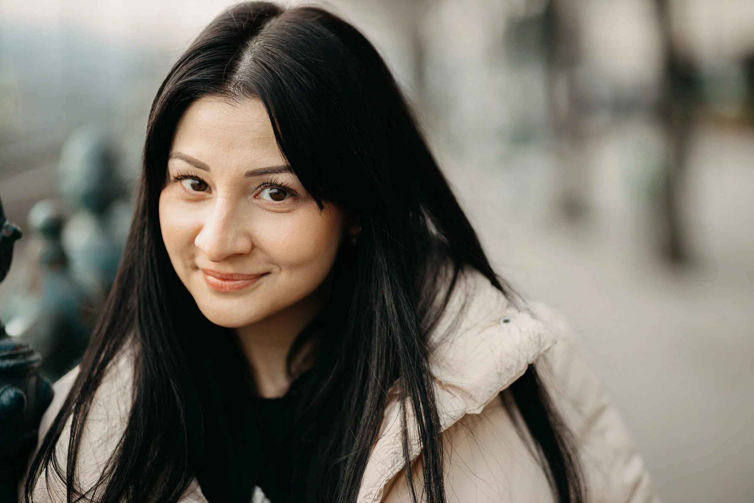 A young woman with long black hair and light skin smiling at the camera, posed outdoors near a bicycle with a blurred background.