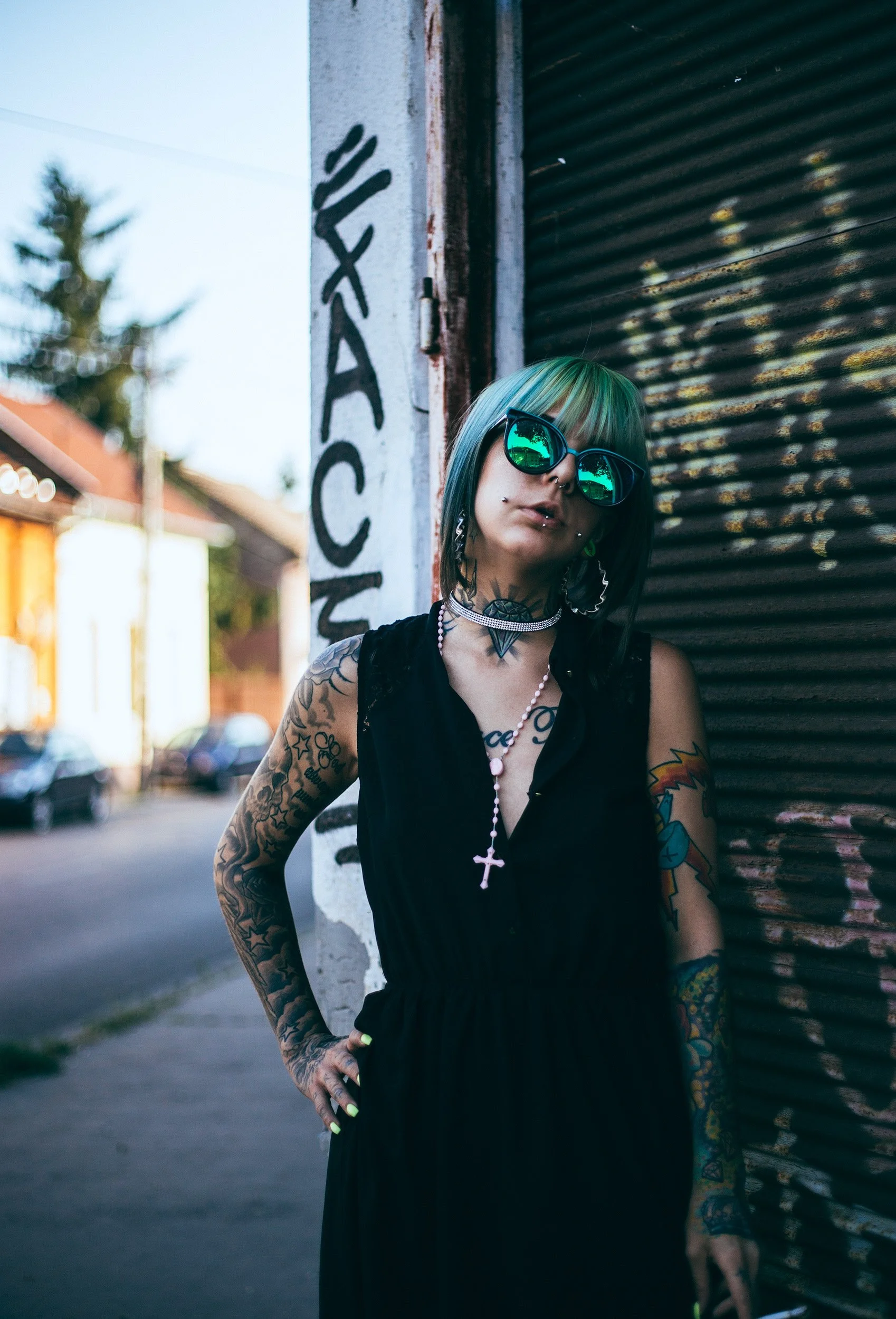 A woman with short multicolored hair and tattoos wearing sunglasses, necklaces, and a black sleeveless dress, standing outdoors next to a rusty metal wall with graffiti, on a street with blurred trees and houses in the background.