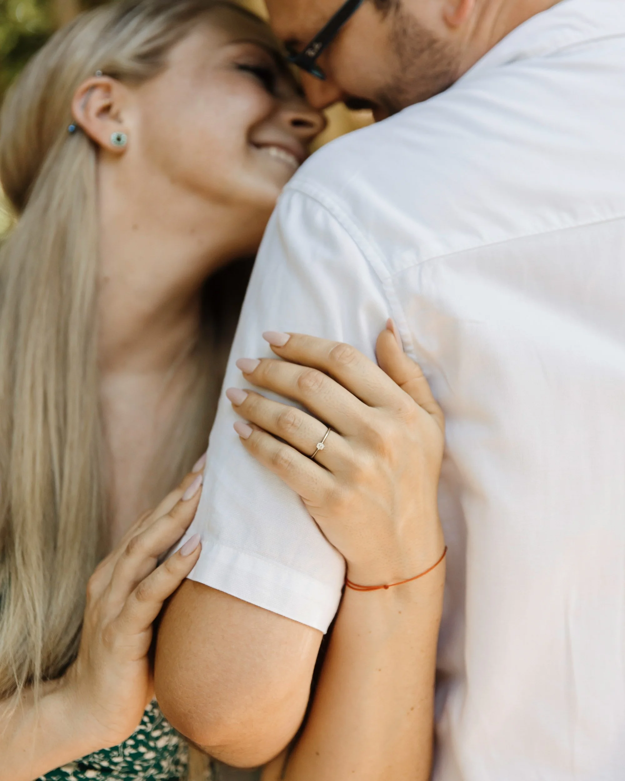 A couple with their eyes closed and foreheads touching, embracing closely. The woman has long blonde hair, earrings, and is wearing a patterned top. The man is wearing glasses, a white shirt, and has a red string bracelet on his wrist.