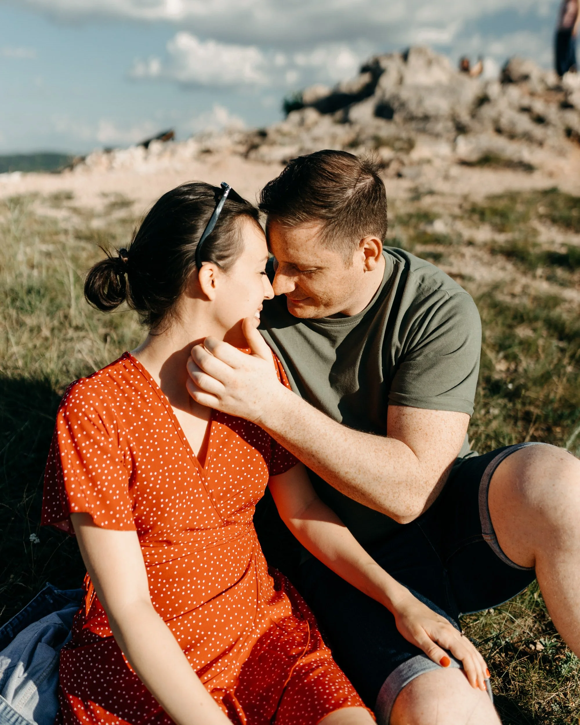 A couple is sitting outdoors on grass with rocky hills and cloudy sky in the background. They are touching foreheads and smiling, with the woman wearing a red polka dot dress and sunglasses on her head, and the man in a green t-shirt and black shorts
