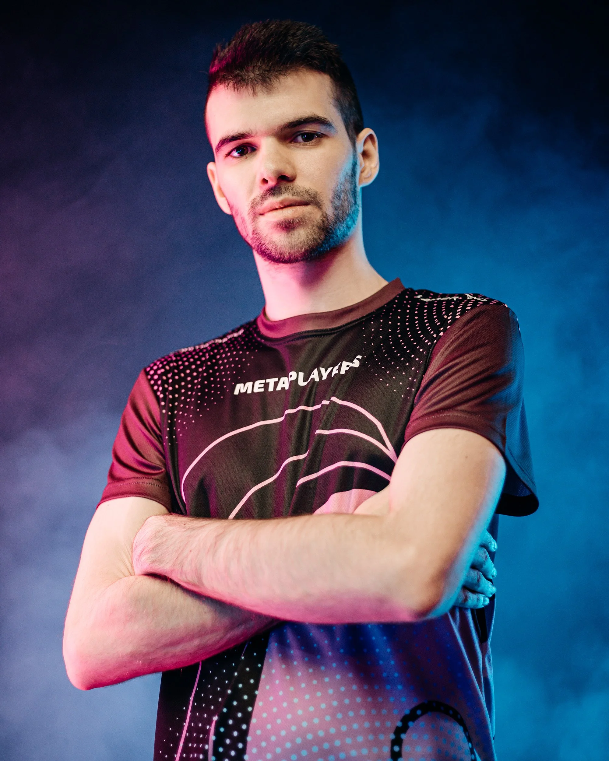 A young man with short dark hair, a beard, wearing a black esports jersey with pink and white accents, standing with crossed arms against a dark background illuminated with pink and blue lighting.