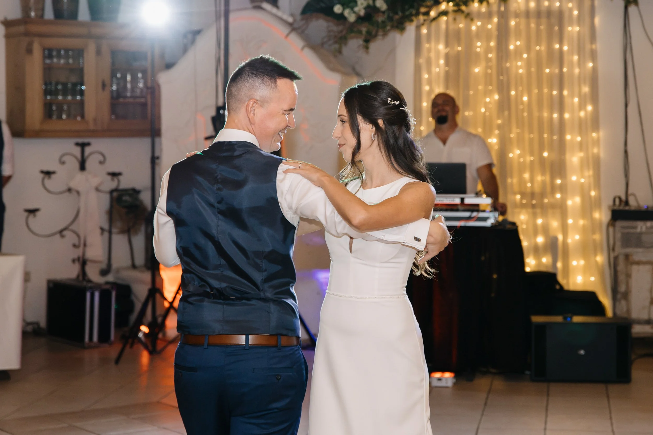 A couple dancing closely at a celebration, with warm lighting and a DJ in the background.