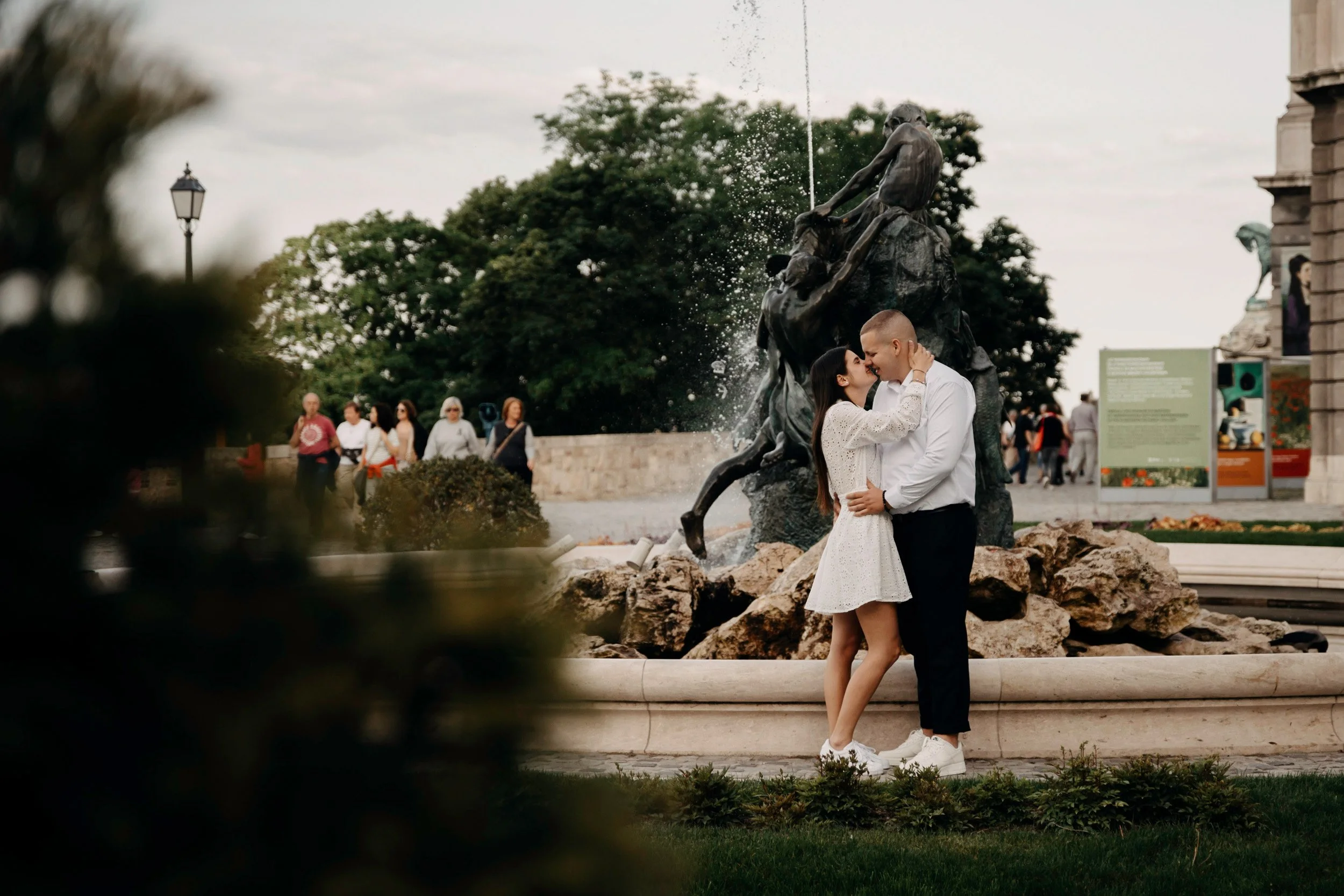 A couple is standing close together and smiling in front of a fountain sculpture in a public park. The woman is wearing a white dress and the man is wearing a white shirt and dark pants. There are trees and several people in the background.