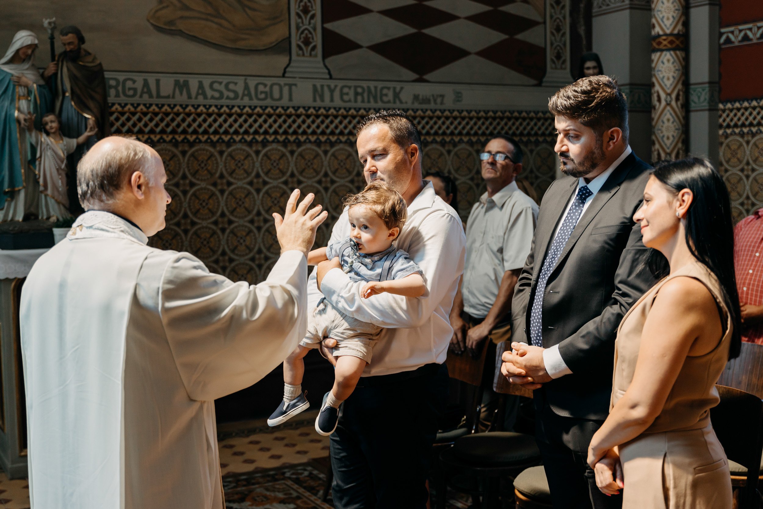 Priest performing a religious ritual or blessing in a church, with people standing in reverence. A man holding a child stands before him, and others, including a woman, are witnessing the ceremony.