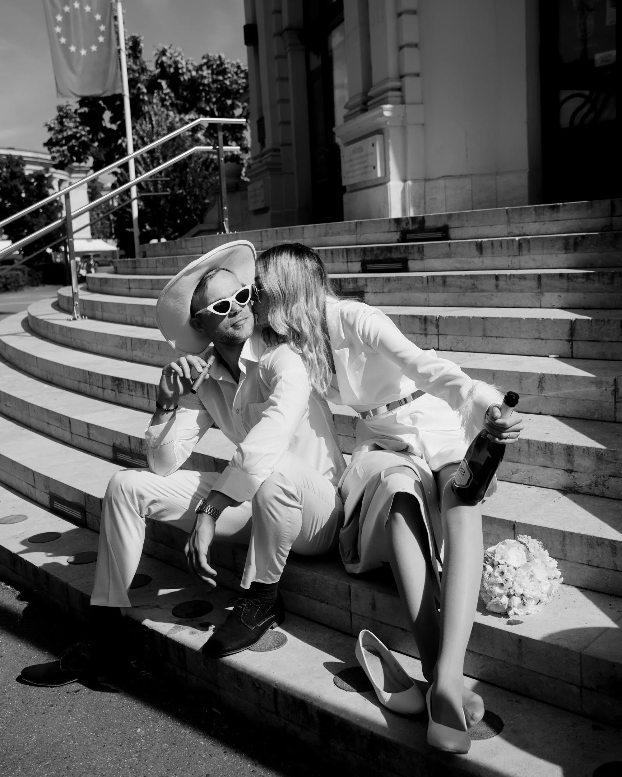 Two people in white clothing sitting on stairs outside a building, one with a hat and sunglasses, holding a bottle of champagne, with flowers on the ground nearby.