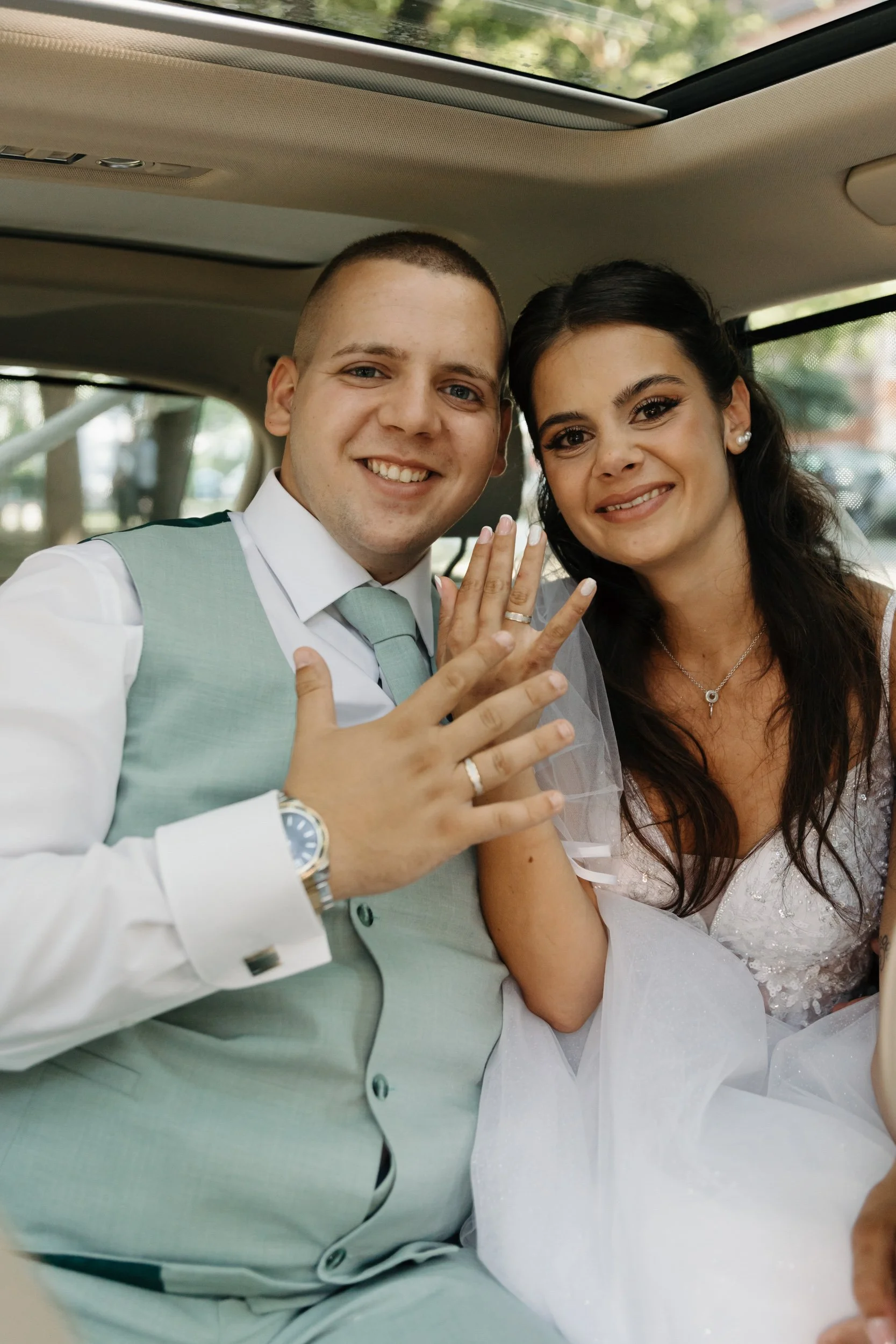 A smiling couple is seated inside a vehicle, with the man showing an engagement ring on his finger and the woman holding his hand, both dressed formally for a special occasion.