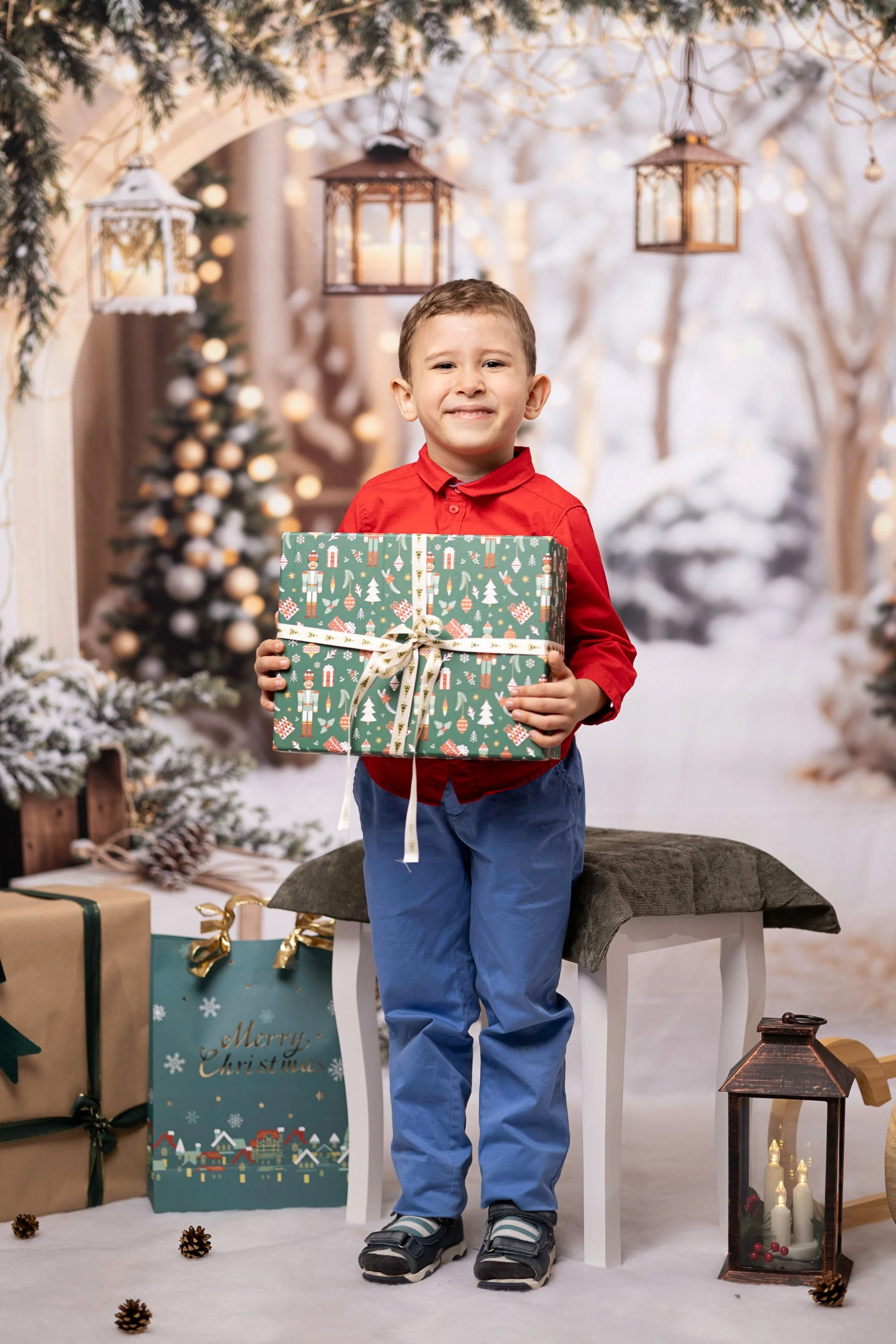 A young boy in a red shirt and blue pants holding a Christmas present, standing in a festive winter scene with snow-covered trees, holiday decorations, and wrapped gifts.