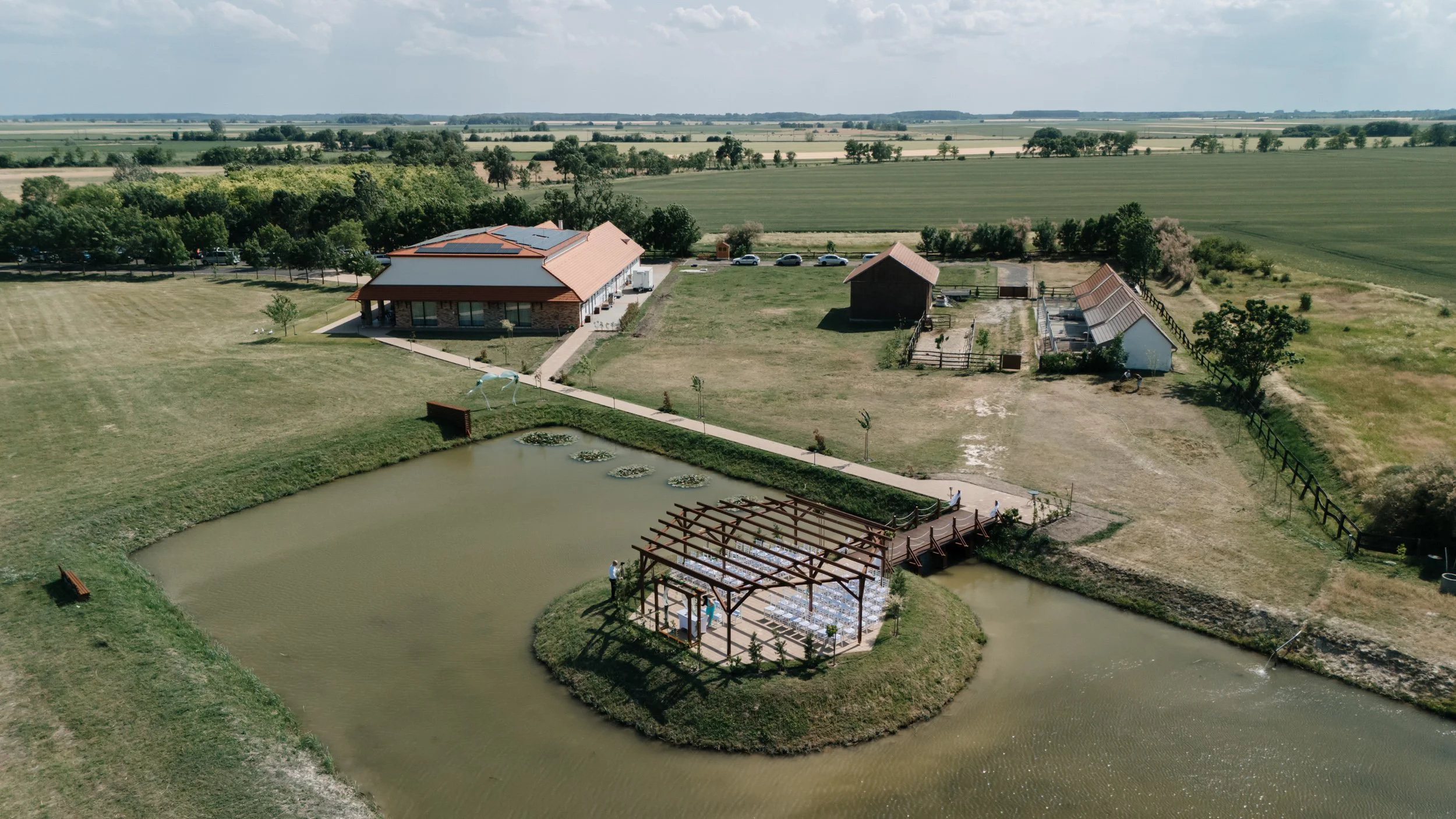 Aerial view of a rural property with a pond, a building with solar panels, a smaller shed, and outdoor seating area with a pergola, surrounded by open fields and farmland.