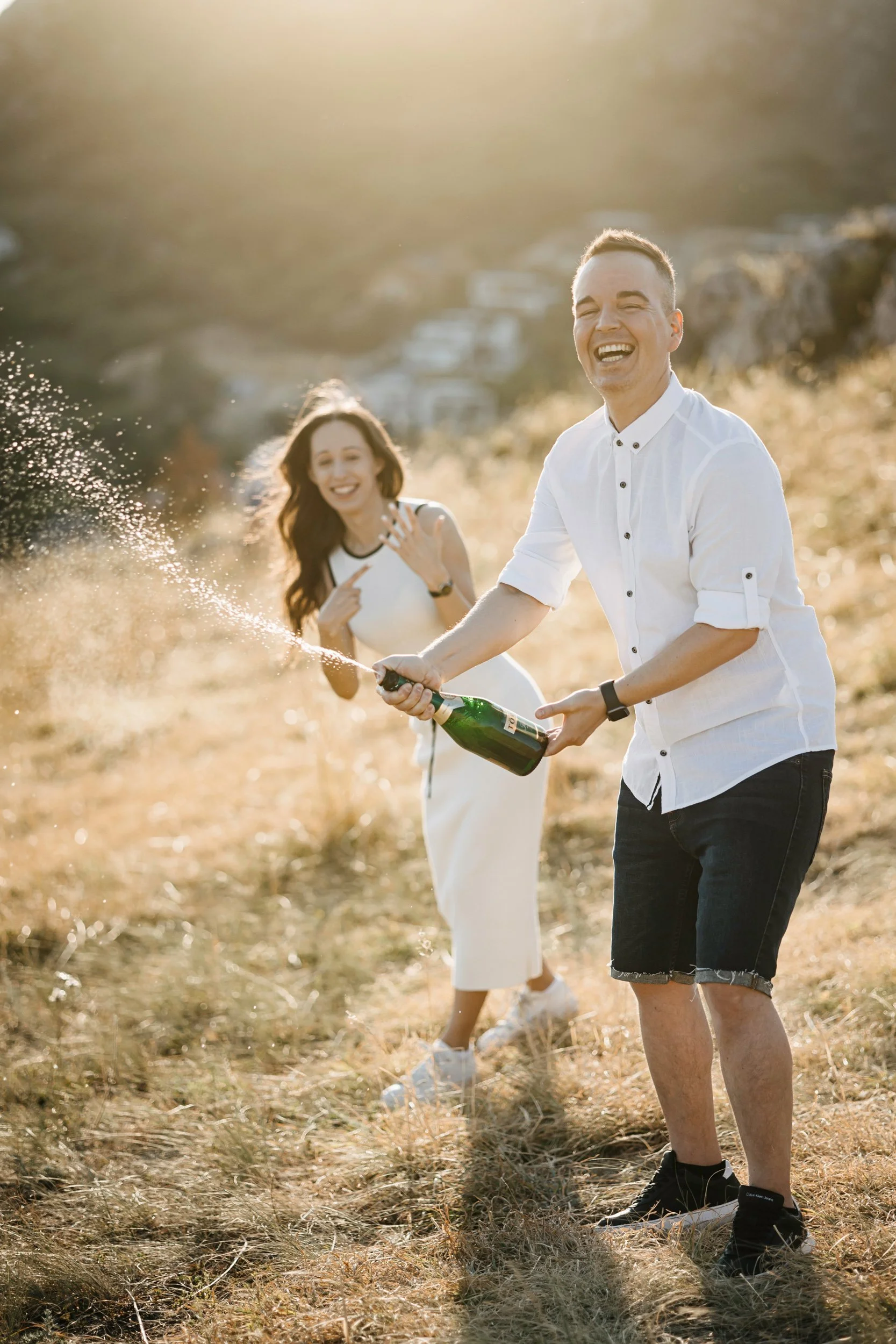 Two friends celebrating outdoors, one holding a champagne bottle and spraying it, the other smiling and pointing, during sunset in a grassy field.