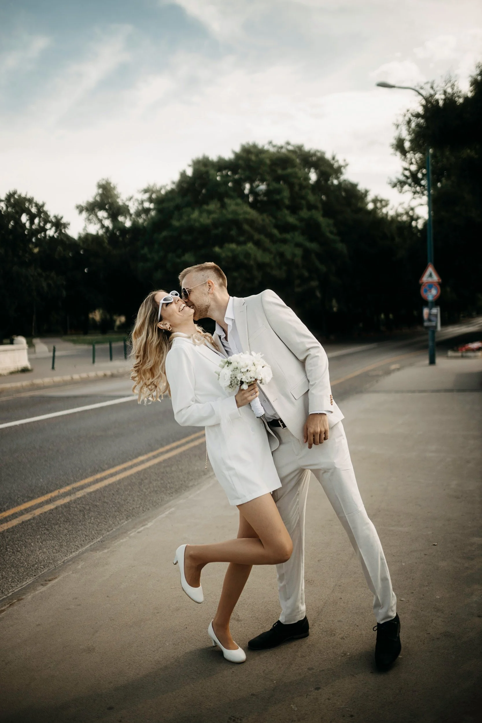 A couple dressed in white clothes sharing a kiss on a street with trees in the background.