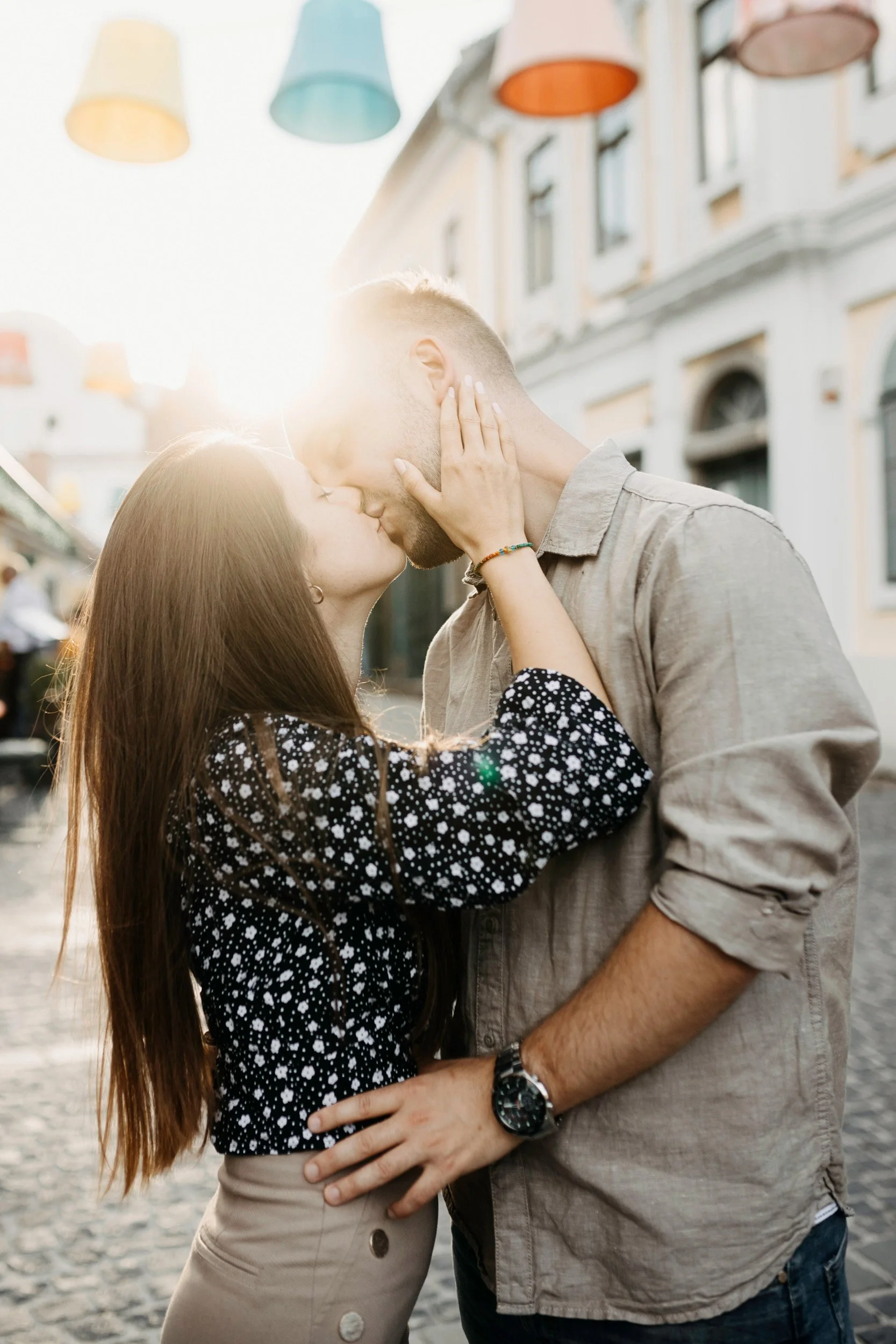A couple sharing a kiss outdoors on a sunny day, with colorful lamps hanging overhead and historic buildings in the background.
