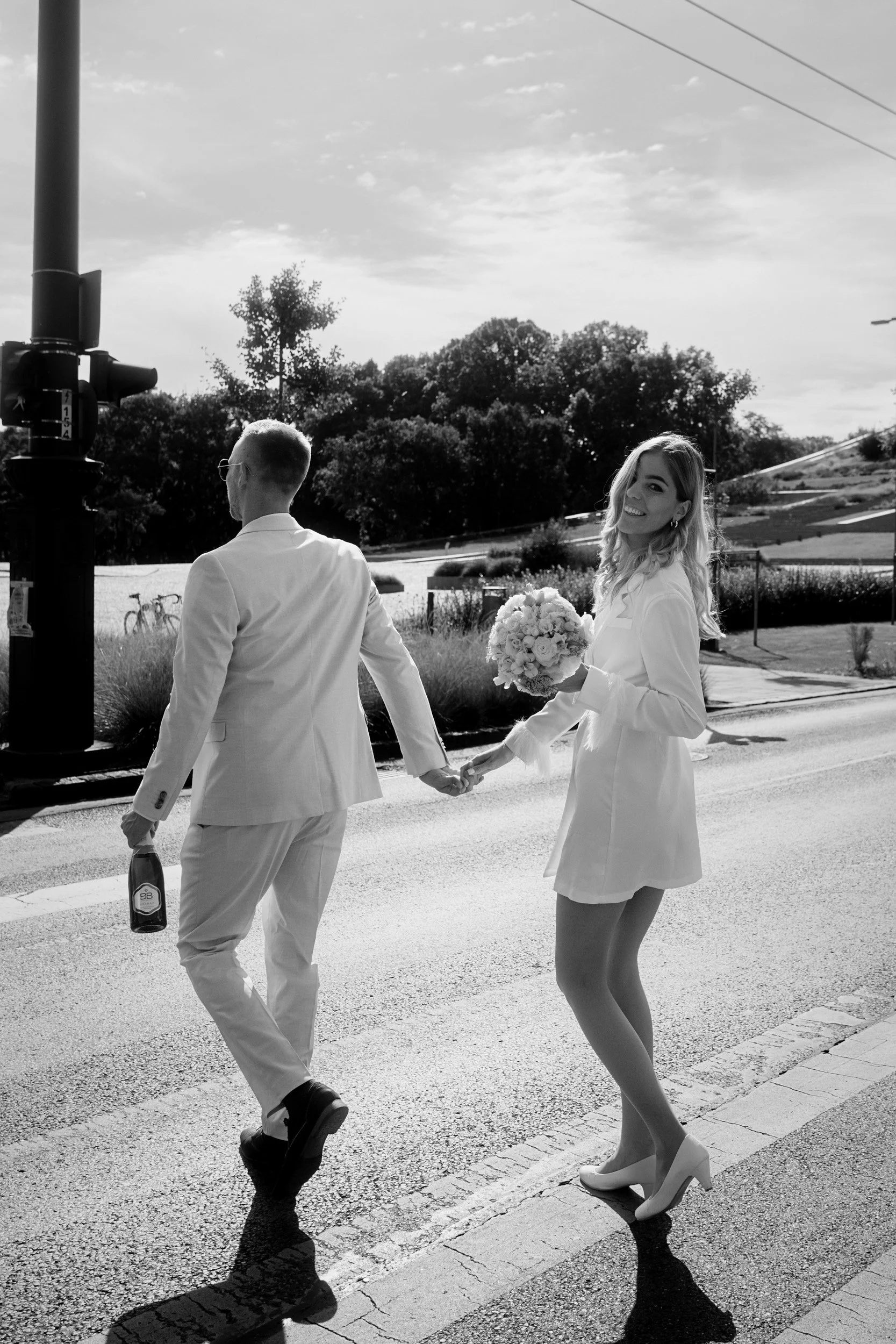 A woman in a white dress with high heels holding a bouquet of flowers, and a man in a light-colored suit holding a bottle, walking hand in hand outdoors near a traffic light, with trees and rolling hills in the background.