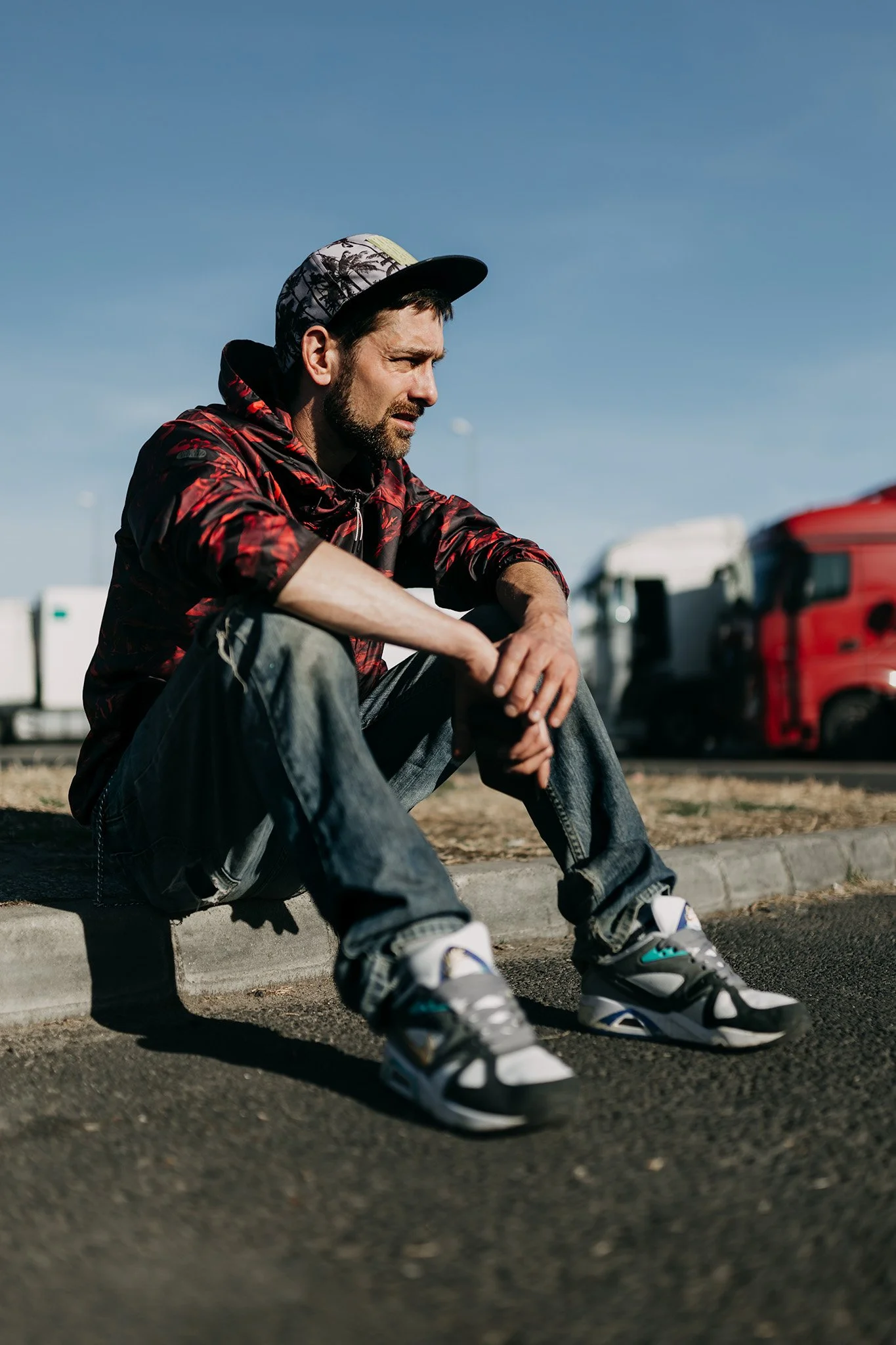 A man sitting on the curb of a parking lot with his head slightly bowed, wearing a patterned jacket, jeans, sneakers, and a baseball cap. Red trucks are parked in the background under a clear blue sky.