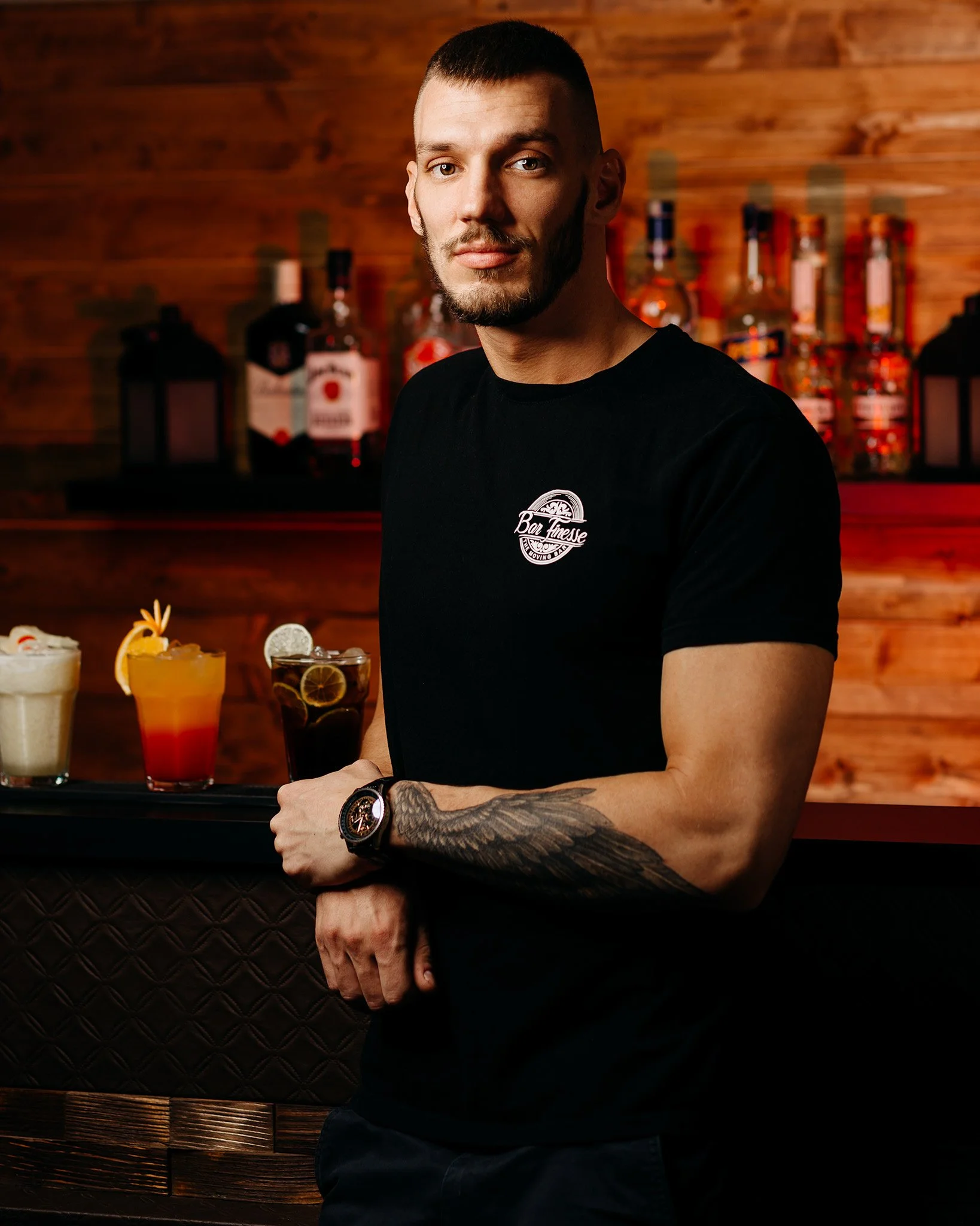 A young man with short hair, beard, and light skin stands in front of a bar with bottles of alcohol on shelves behind him. He is wearing a black t-shirt with a logo, a wristwatch, and has tattoos on his arm. The background features wooden paneling an