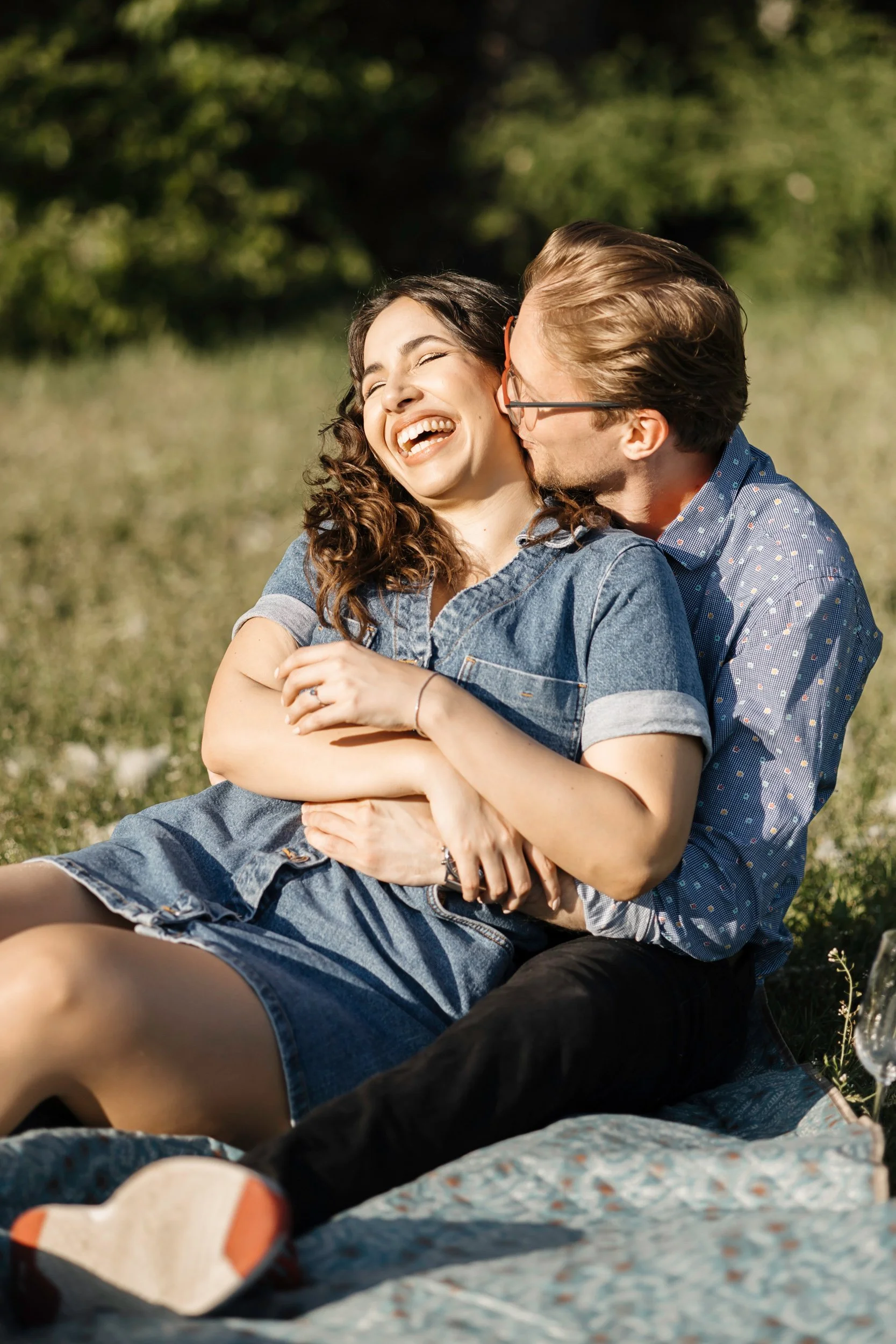 A young woman with curly hair laughing and embracing a young man with glasses and slicked-back hair, both sitting outdoors on grass during daytime.