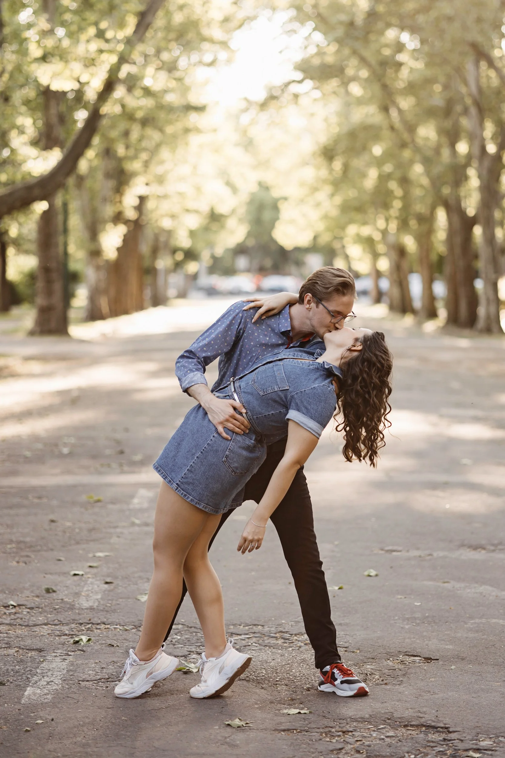 A couple dancing outdoors on a tree-lined street during the day, with the man dipping the woman and leaning in for a kiss.