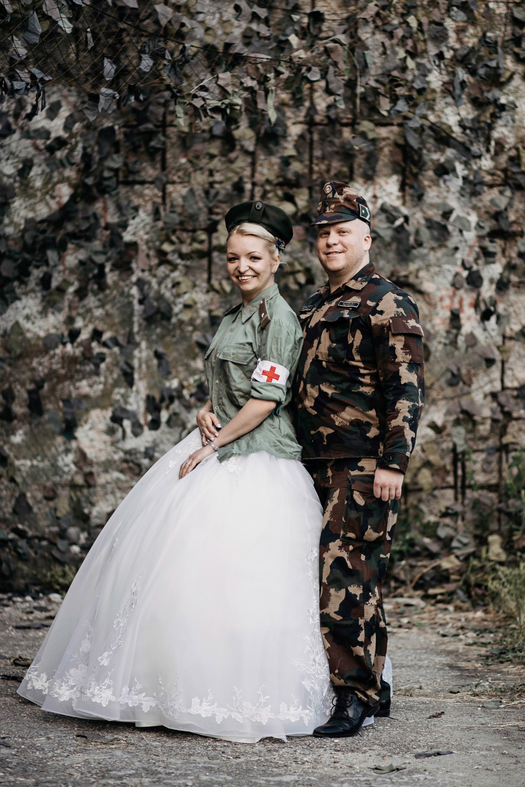 A couple dressed in military uniforms, with the woman in a white wedding dress, standing outdoors in front of a rocky background, smiling at the camera.